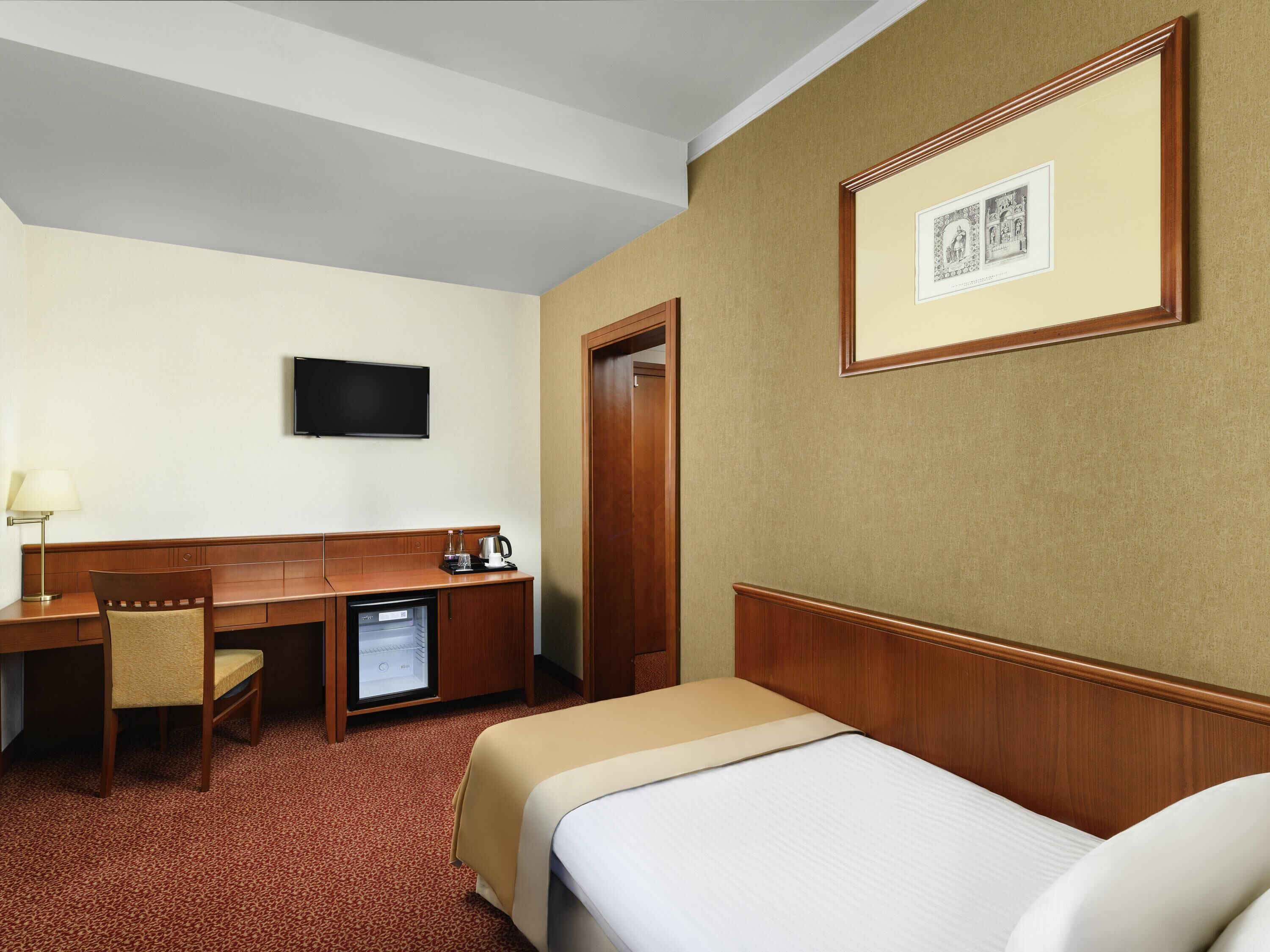 A hotel room with a bed, a desk with a chair, a television, and a small refrigerator.