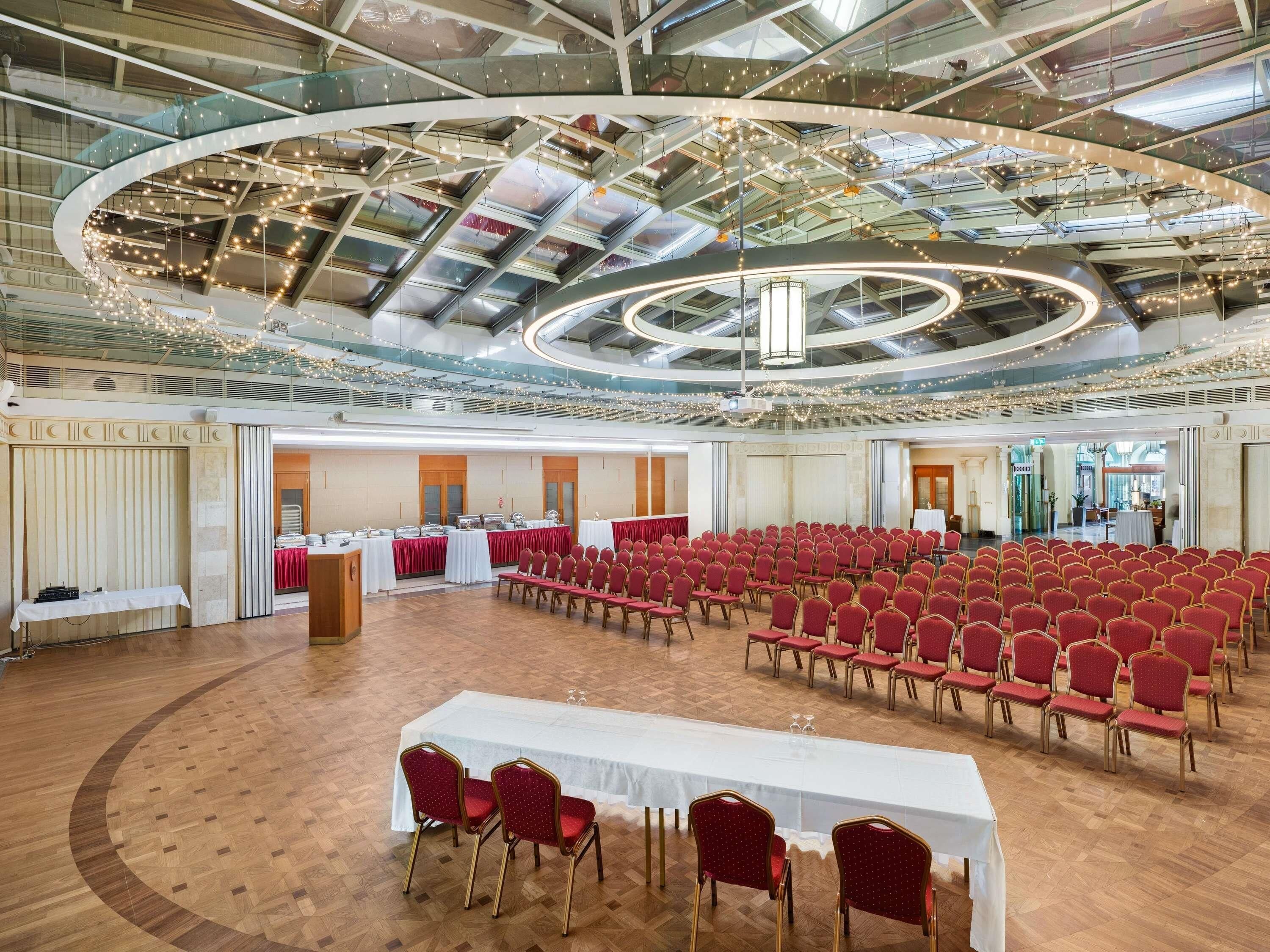 Un salón amplio con techo alto, varias mesas y filas de sillas.
