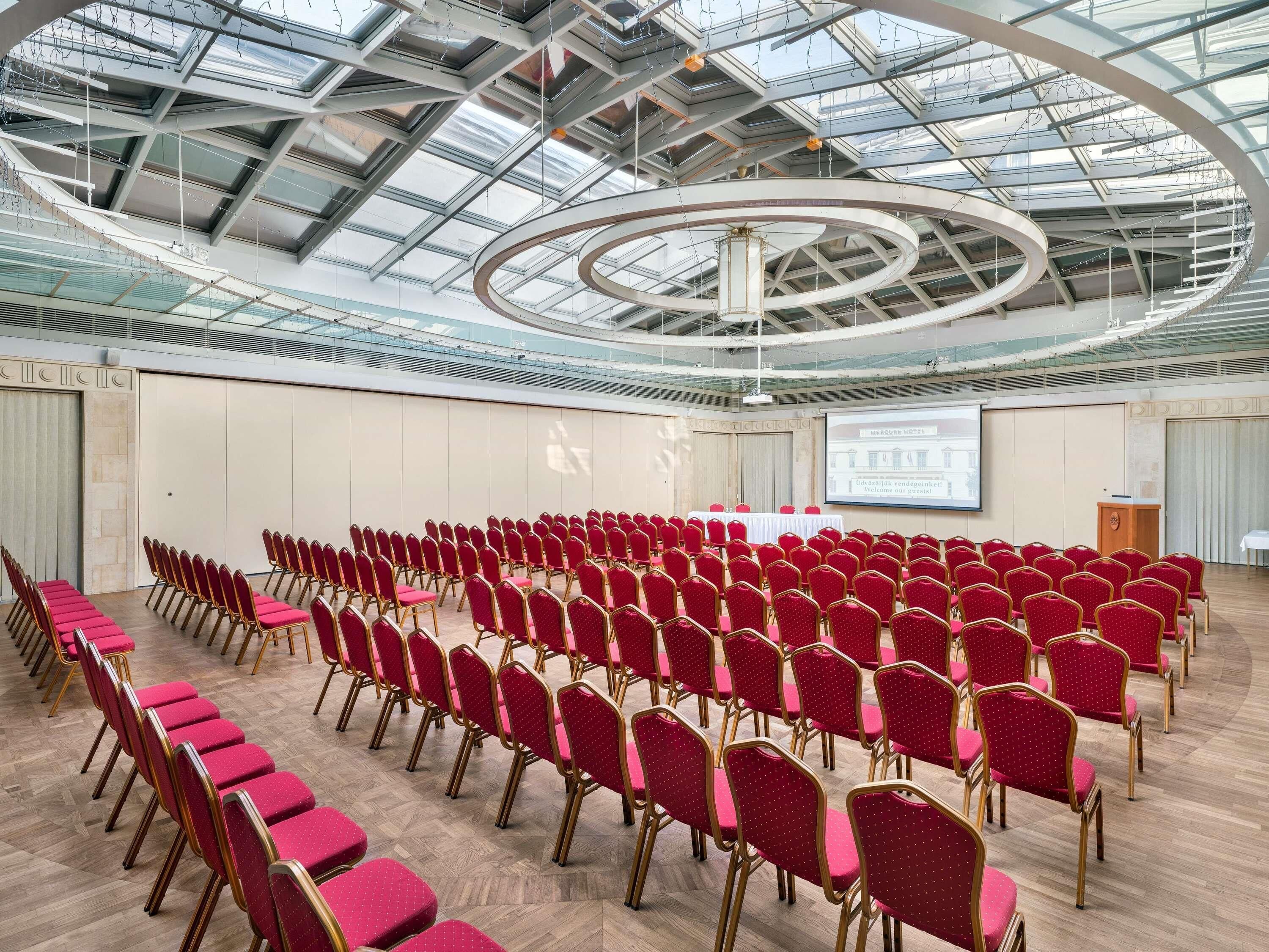 Un salón de conferencias con hileras de sillas rojas y una pantalla grande mostrando un mensaje de bienvenida.