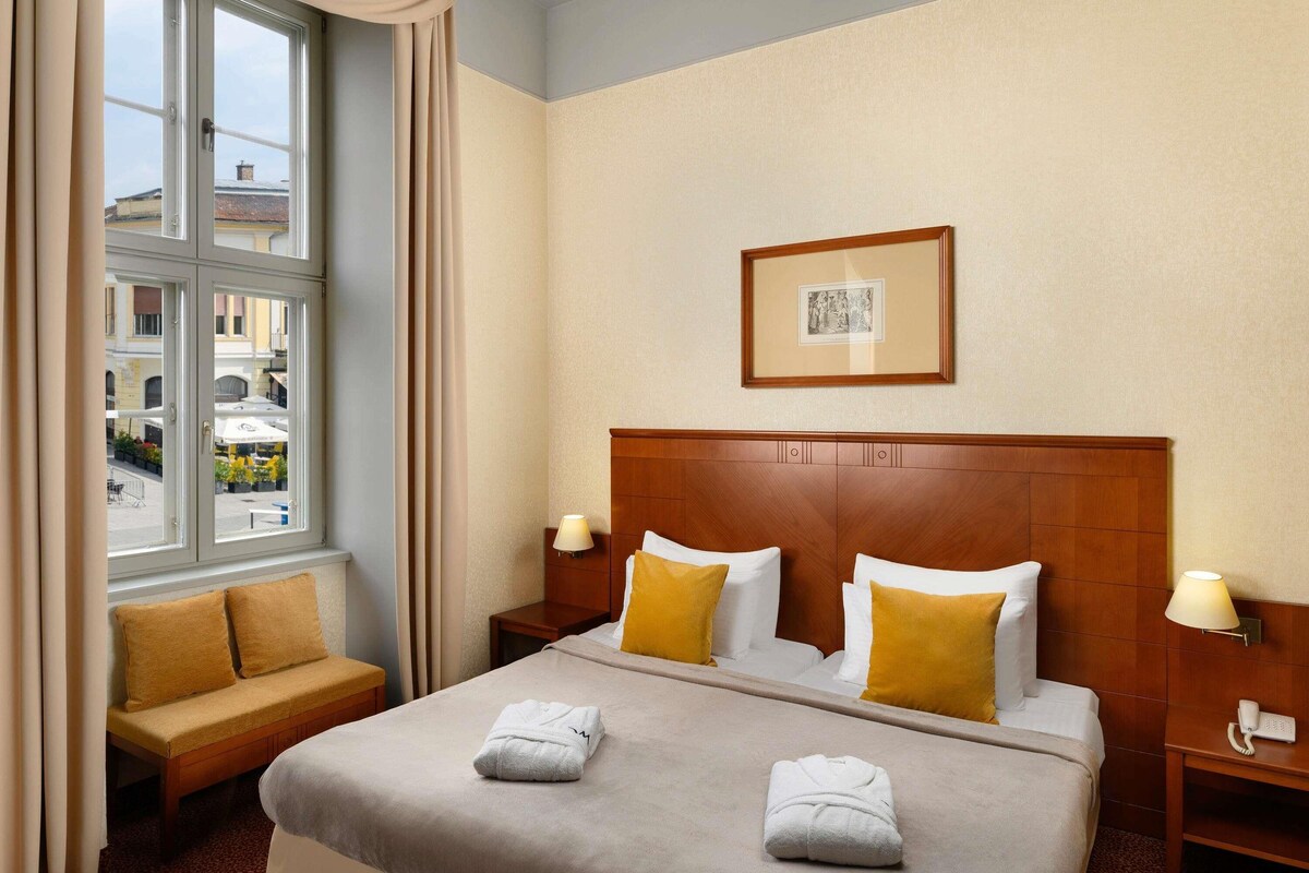 A hotel room with a large bed, two bedside tables with lamps, a wooden headboard, a framed picture on the wall, and a window with curtains.