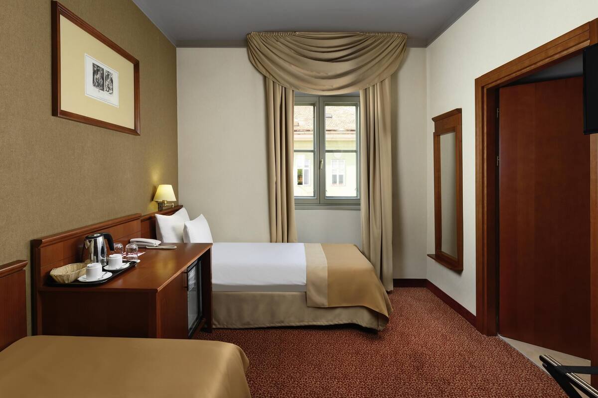 Ein Hotelzimmer mit Bett, Nachttisch, Lampe, Fenster mit Vorhängen und einem gerahmten Bild an der Wand.