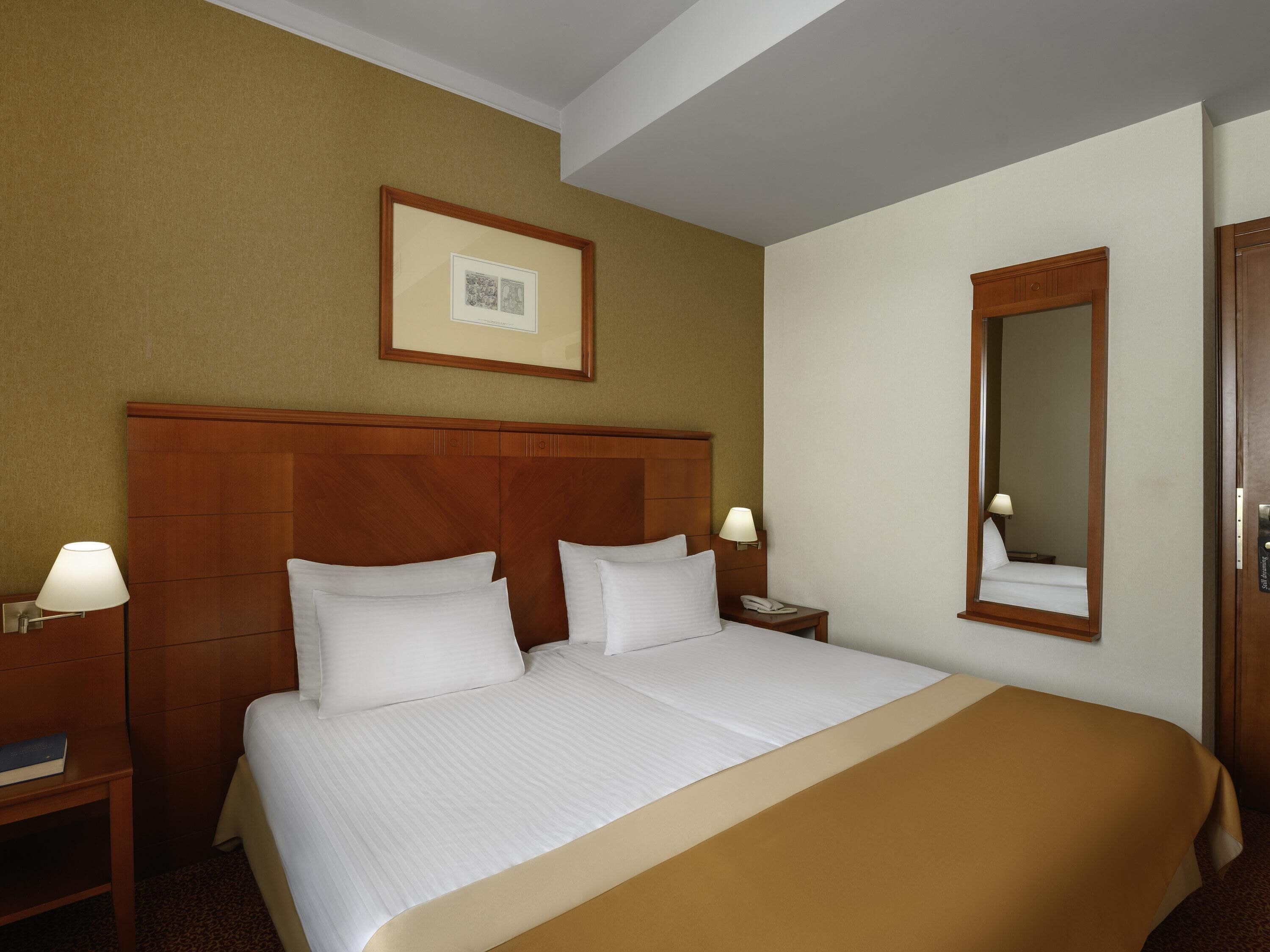 Habitación de hotel con una cama grande, dos mesitas de noche, una mesita auxiliar, un cuadro en la pared y un espejo.