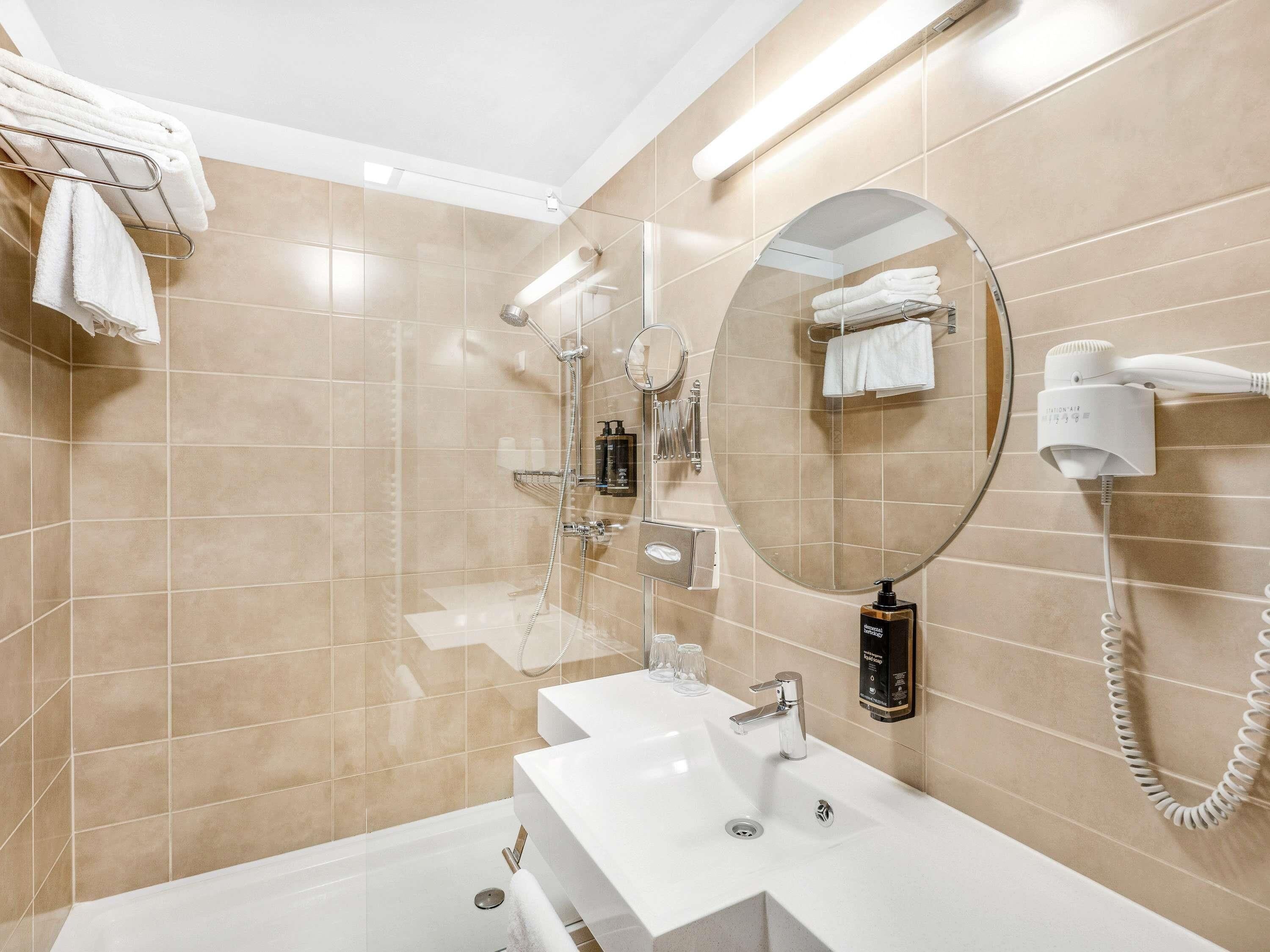 A modern bathroom with beige tiles, a large mirror, a round mirror, a hairdryer, and a towel rack.