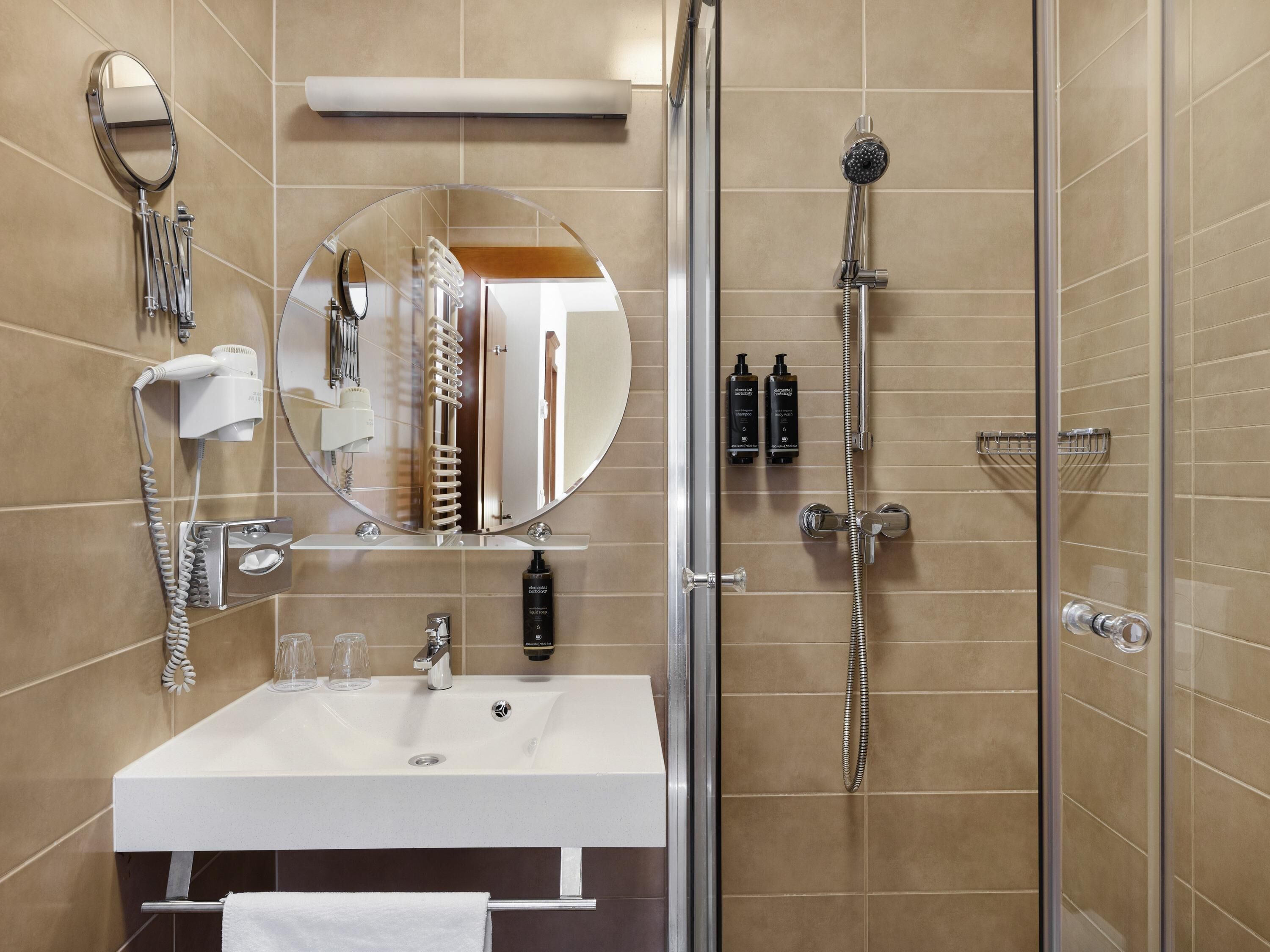 A modern bathroom with a white sink, a round mirror, a handheld showerhead, and a hairdryer.
