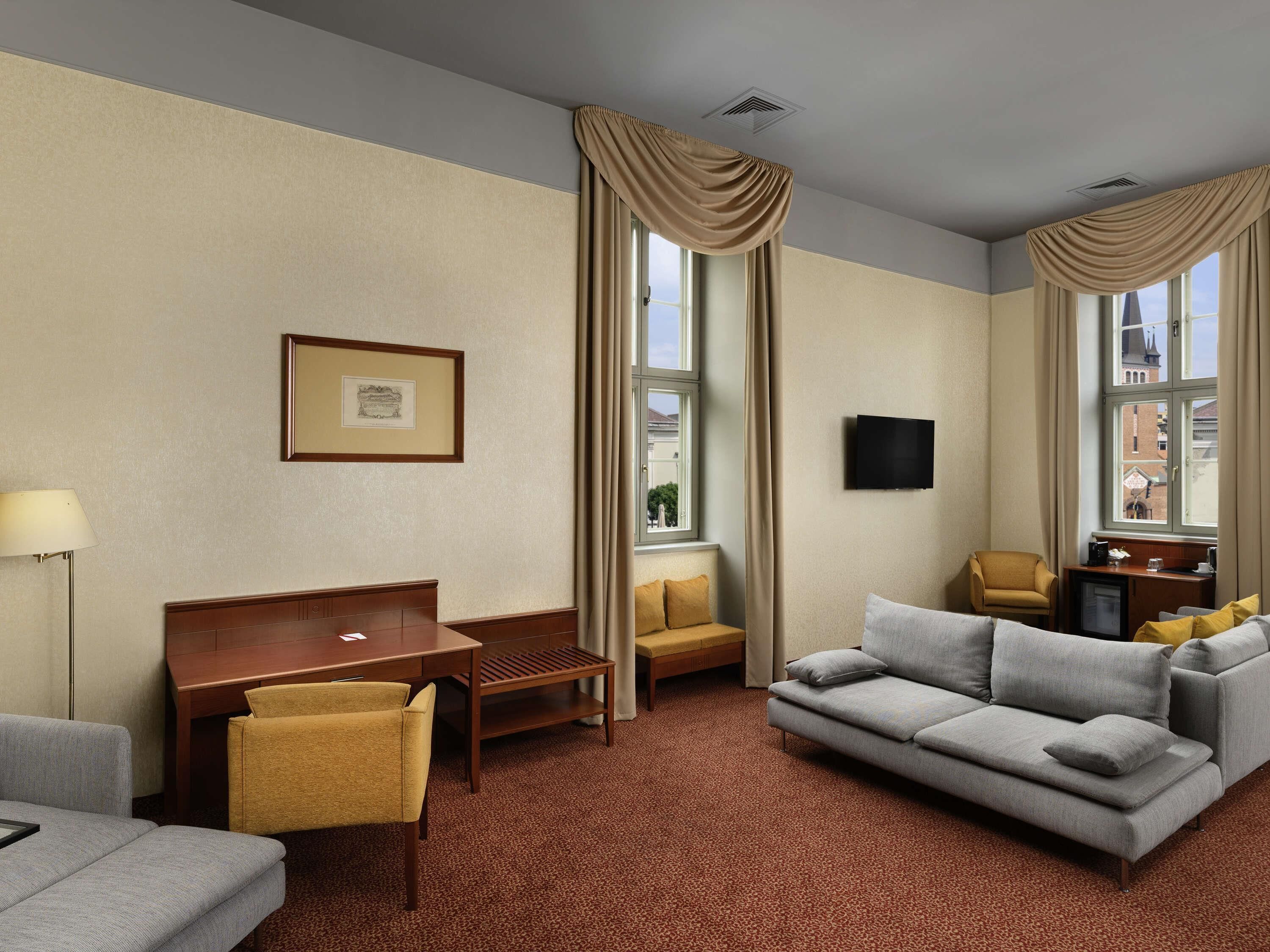 A hotel room with a sofa, a small table, a chair, a desk, a television, and a window with curtains.