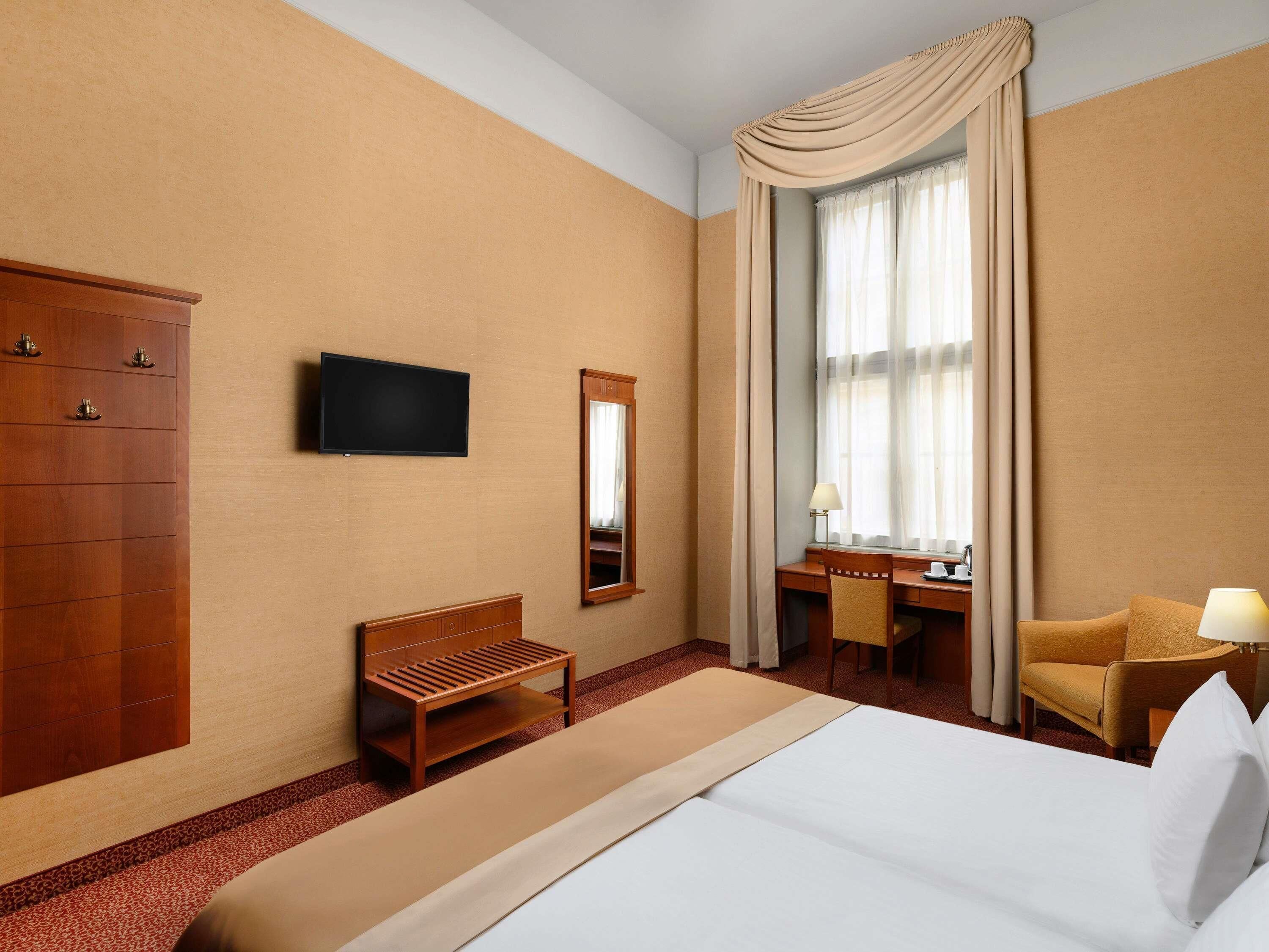 A hotel room with a bed, a desk, a chair, a TV, a wardrobe, and a large window with curtains.