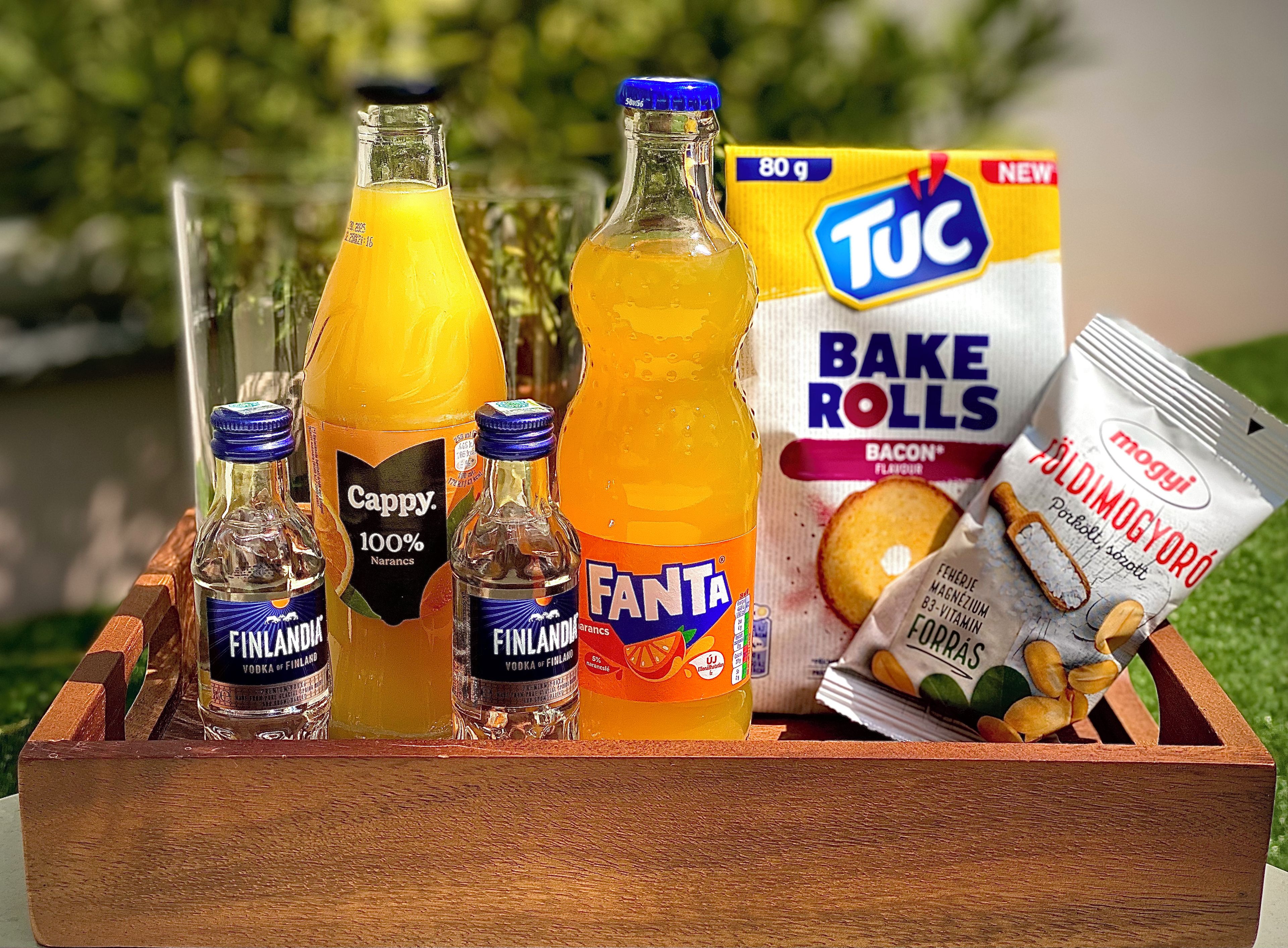 A wooden box containing various food items and beverages, including bottles of Finlandia vodka, a bottle of Fanta, a box of TUC bacon-flavored rolls, and a packet of Mogyoró coffee.