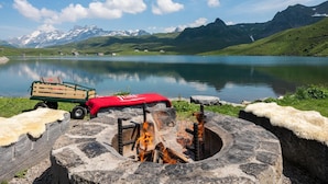 A stone fire pit with logs, a wooden cart, and a red blanket in a mountainous landscape with a lake and snow-capped peaks in the background. - Frutt Mountain Resort (Kerns)