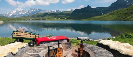A stone fire pit with logs, a wooden cart, and a red blanket in a mountainous landscape with a lake and snow-capped peaks in the background.