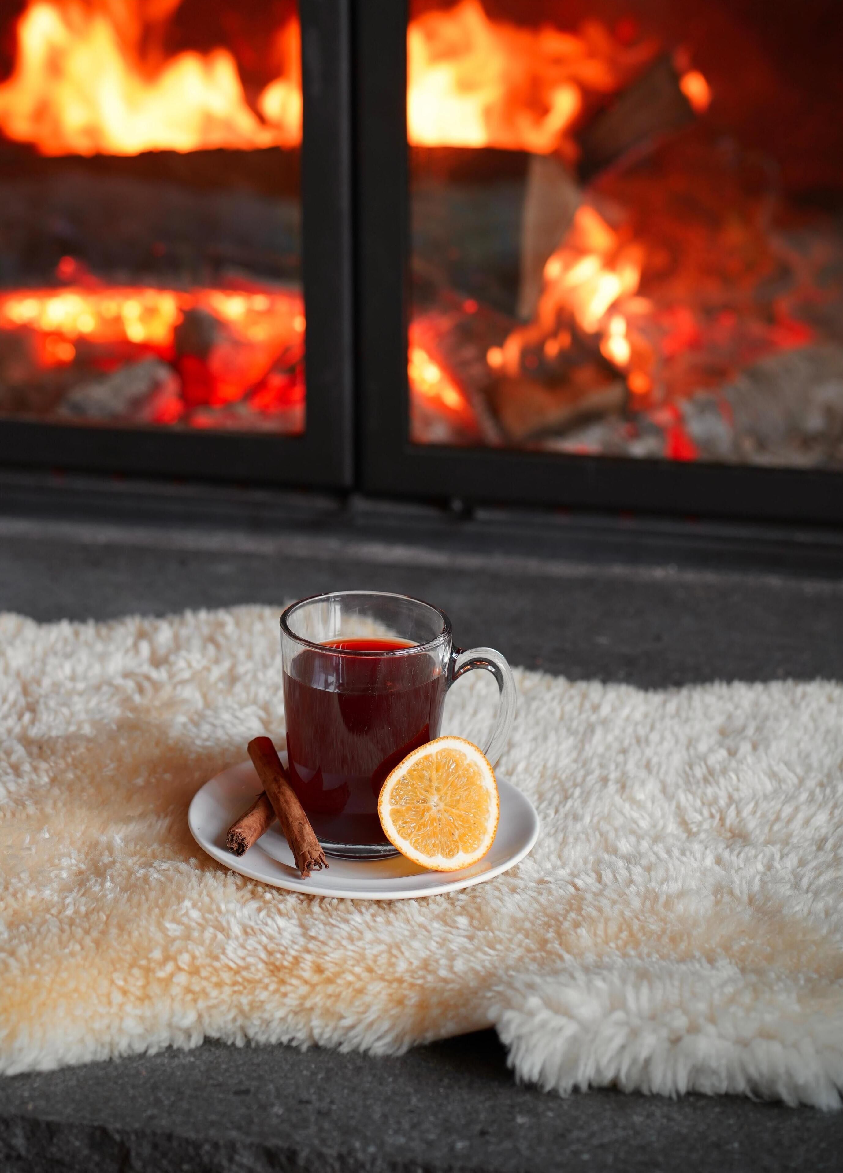 A glass mug of red beverage on a white plate with a slice of orange and cinnamon sticks, set on a fluffy white rug by a fireplace with visible logs.