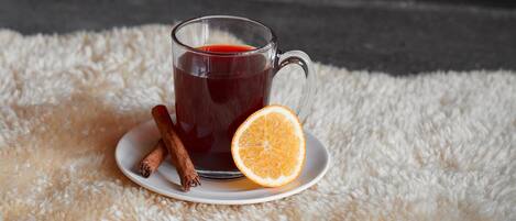 A glass mug of red beverage on a white plate with a slice of orange and cinnamon sticks, set on a fluffy white rug by a fireplace with visible logs.