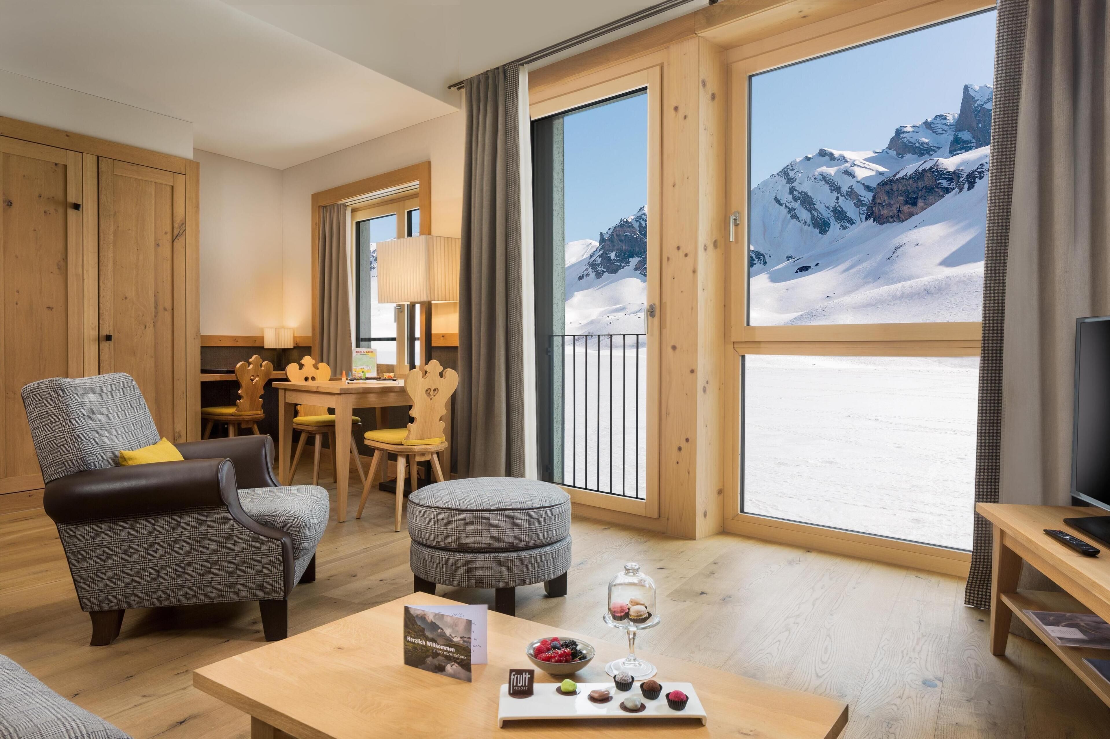 A cozy room with a large window offering a snowy mountain view, a wooden table with a fruit tray, and a comfortable seating area.