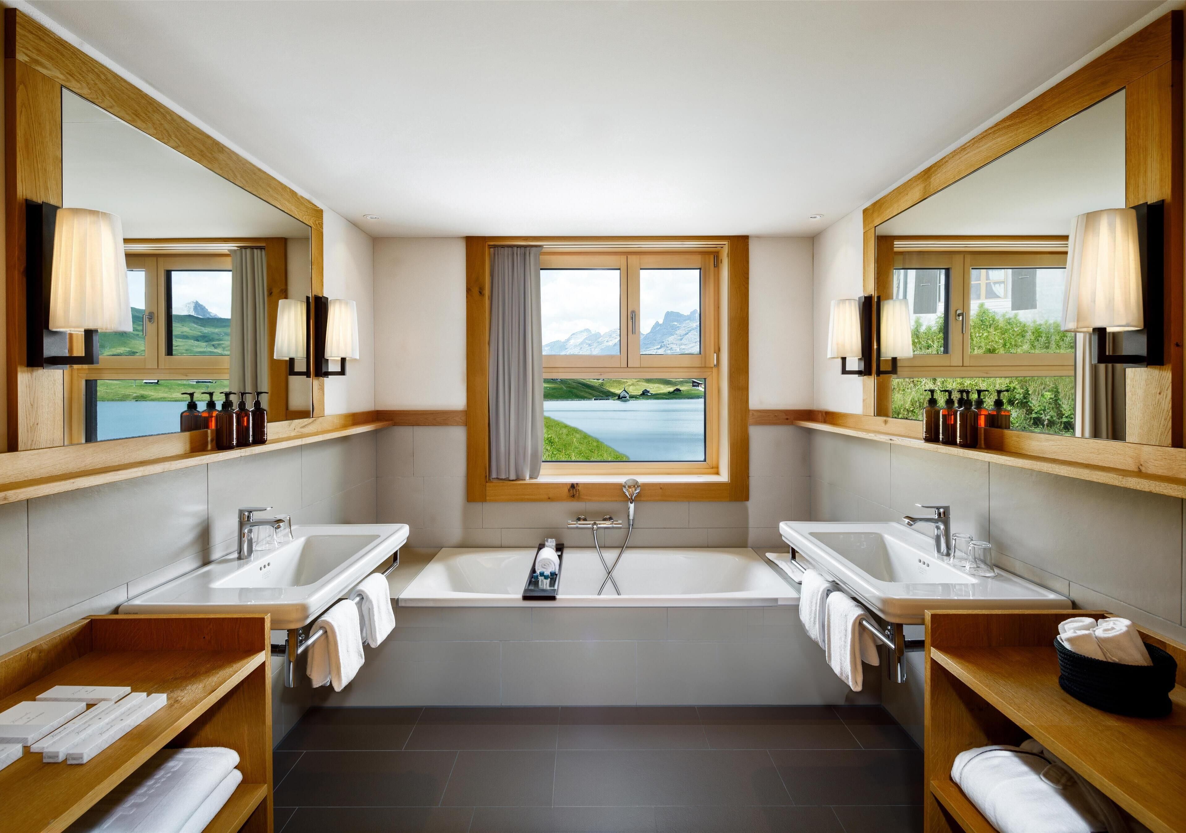 Een moderne badkamer met twee wastafels, een ruime badkuip en een raam met uitzicht op een meer en bergen.