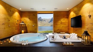 Un cuarto de spa con una bañera grande, un sofá con toallas, un televisor y vistas a un lago y montañas.