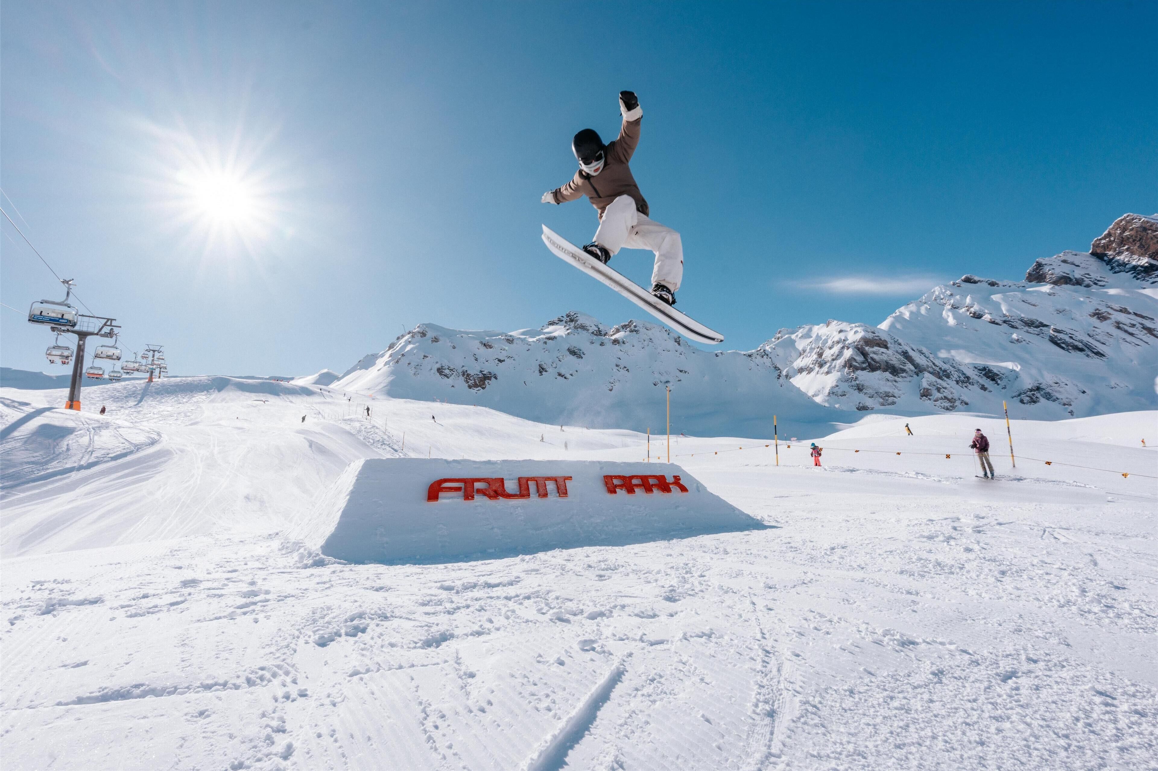 Un snowboarder realizando un salto sobre un letrero que dice 'FRUIT PARK' en una ladera nevada de montaña.