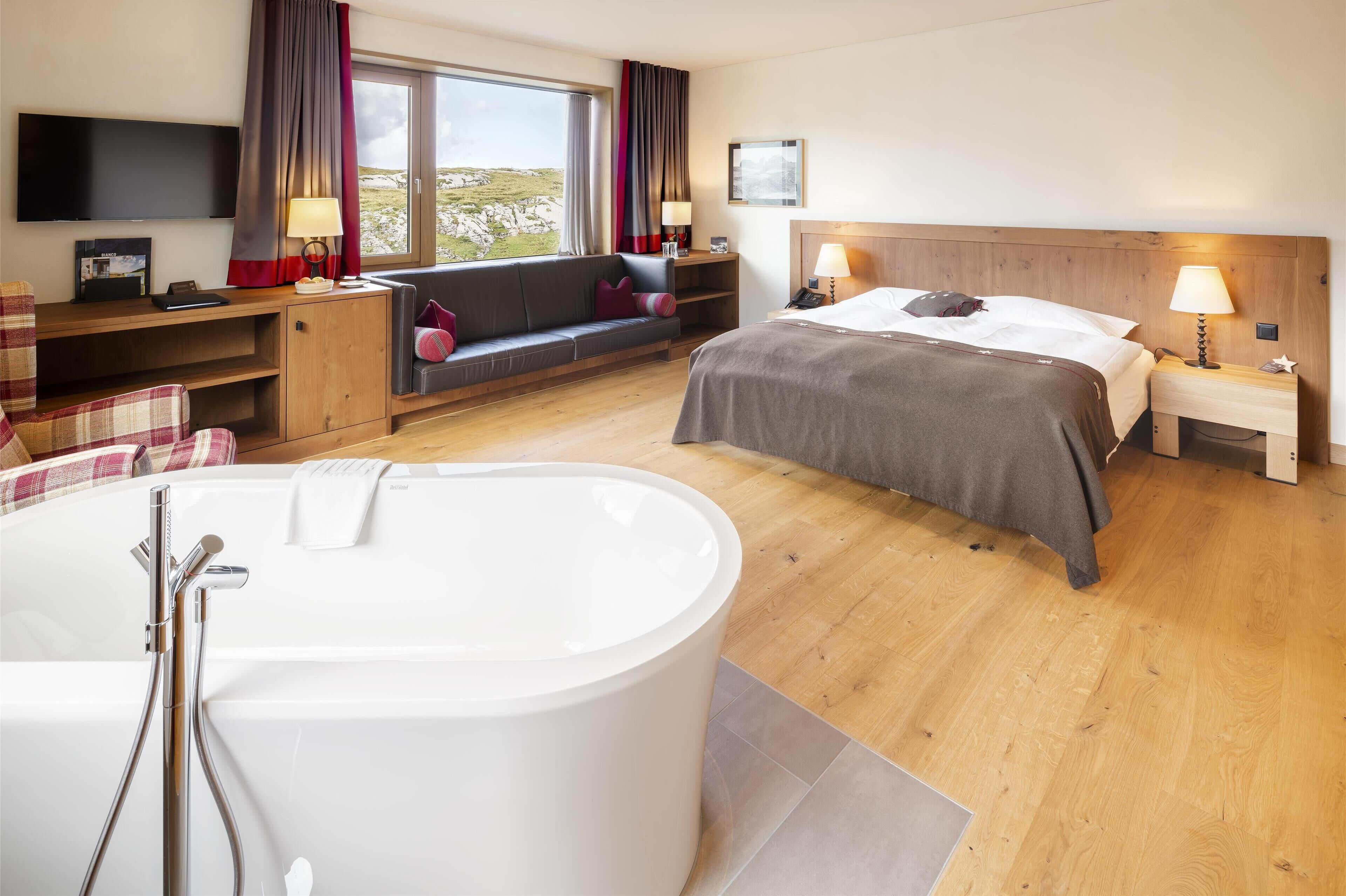 A modern hotel room with a large bathtub, a sofa with a view, a bed with a wooden headboard, and a flat-screen TV mounted on the wall.