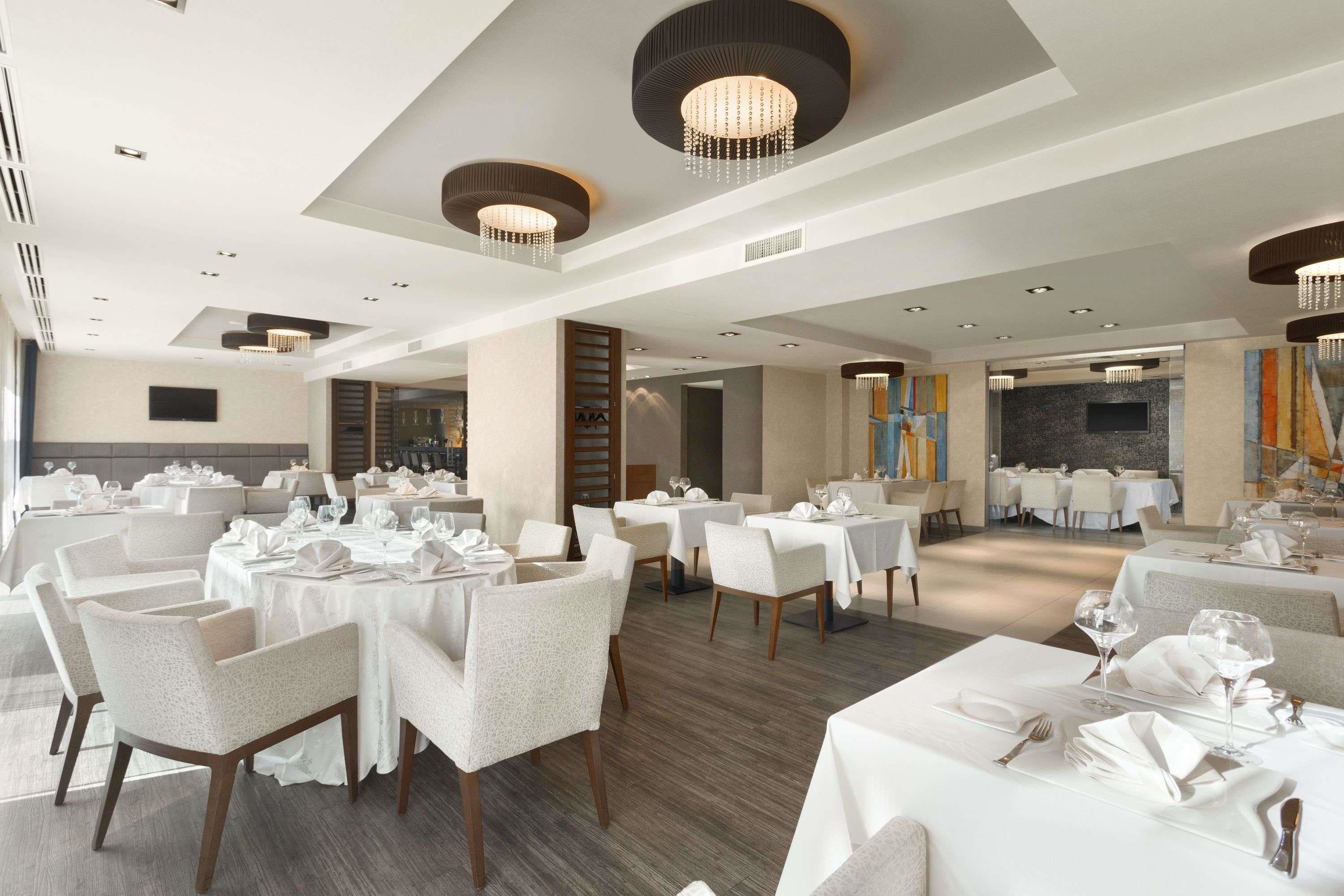 Un comedor amplio con varias mesas dispuestas para recibir a los huéspedes, con candelabros modernos y una paleta de colores neutros.