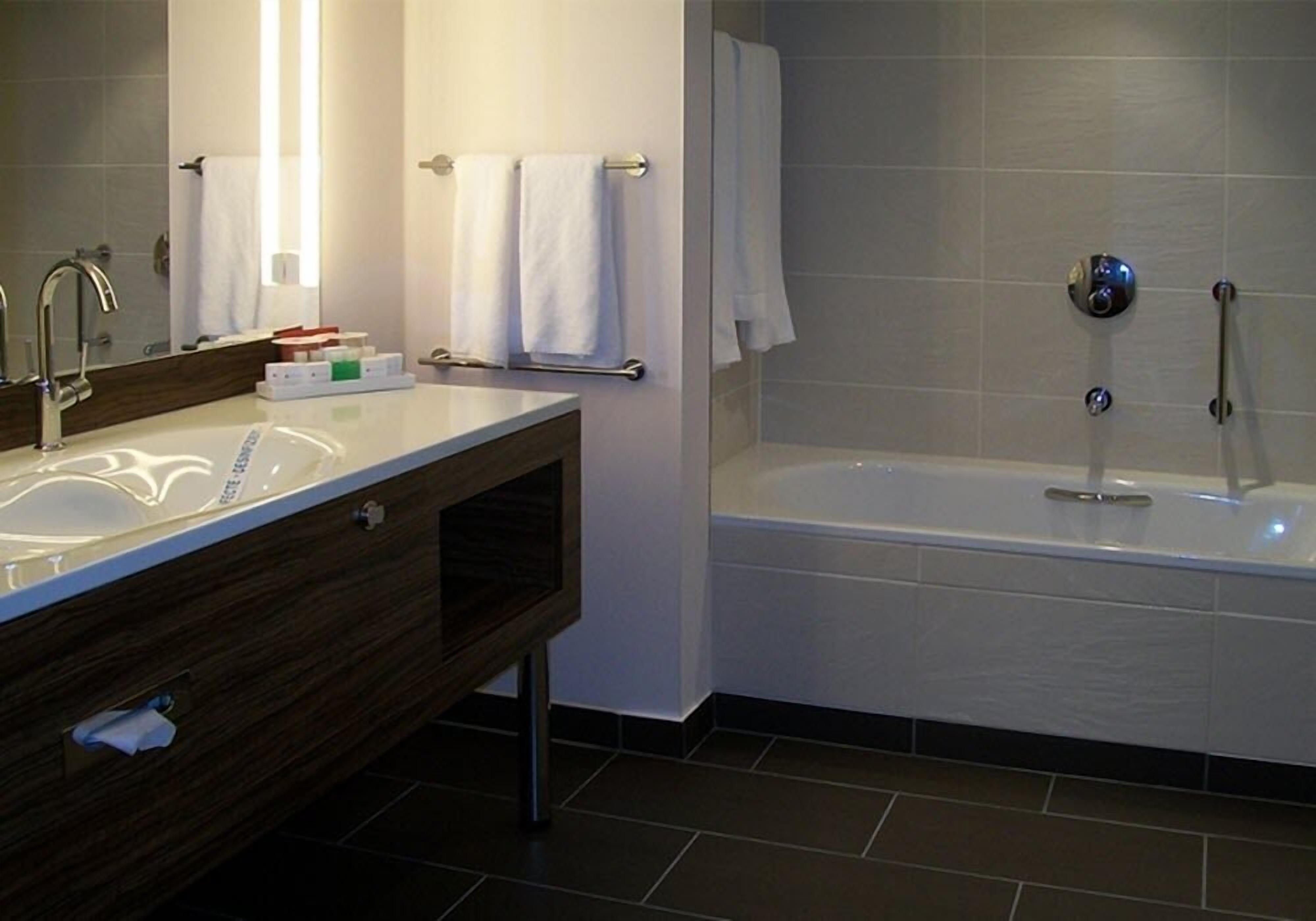 A modern bathroom with a large mirror, a white sink, a vanity with a mirror, a bathtub, and a towel rack.