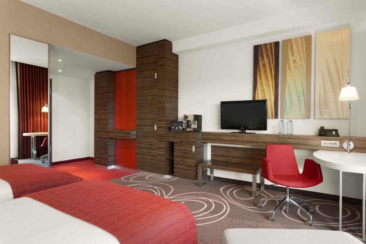 A hotel room with two beds, a desk, a TV, and a red chair.
