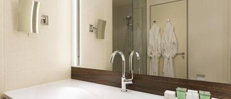 A modern bathroom with a large white sink, two chrome faucets, and a mirror above. There are white robes hanging on a rack and a shower visible in the background.
