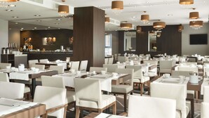 A modern dining area with multiple tables and chairs, large chandeliers, and a bar area in the background. - Ramada by Wyndham Pitesti (Pitesti)