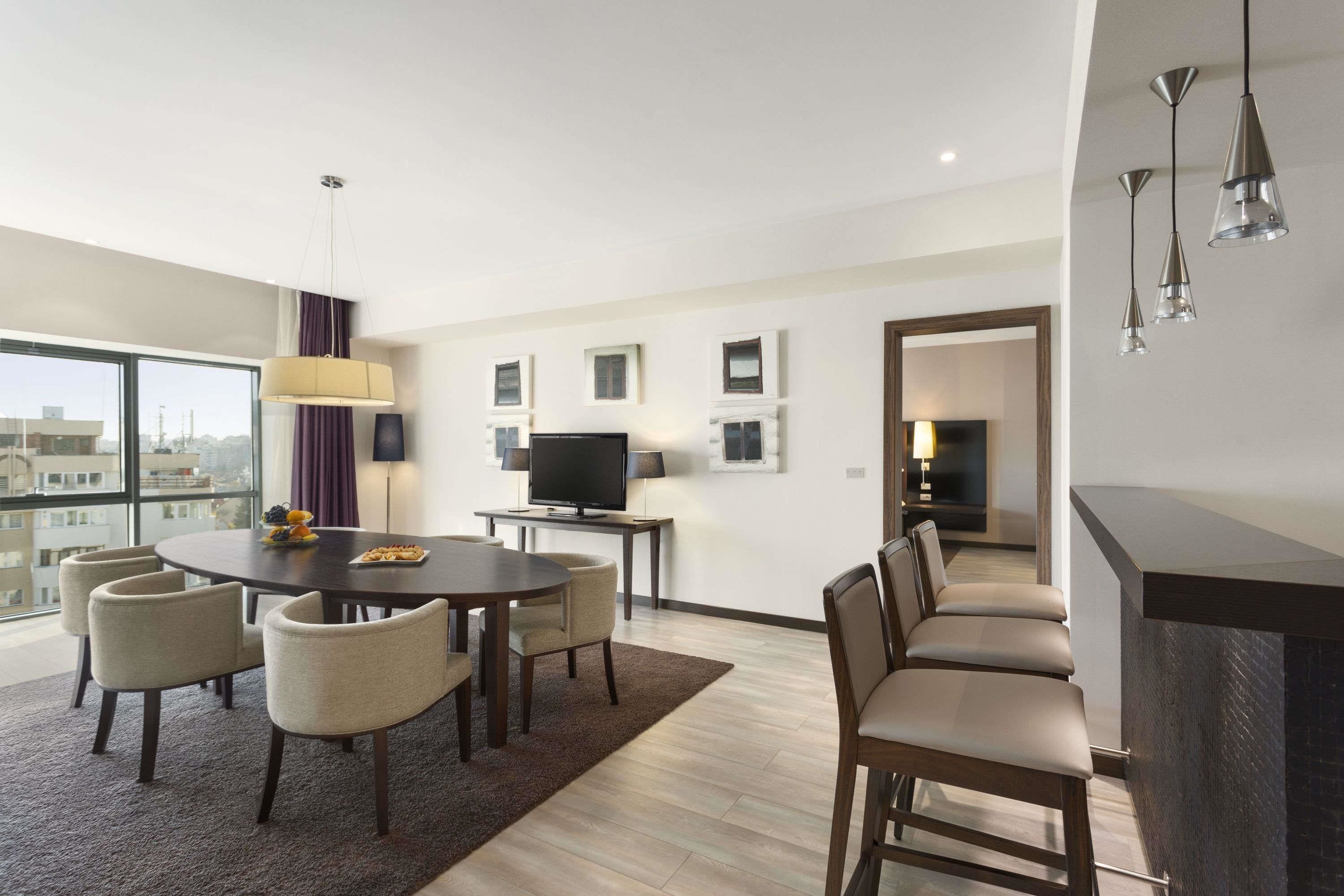 A modern hotel room with a dining area, a TV, and a balcony with a city view.