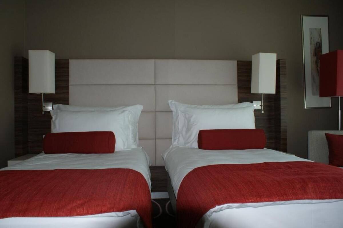 A hotel room with two beds, each with white linens and red accents, and wall-mounted lamps.