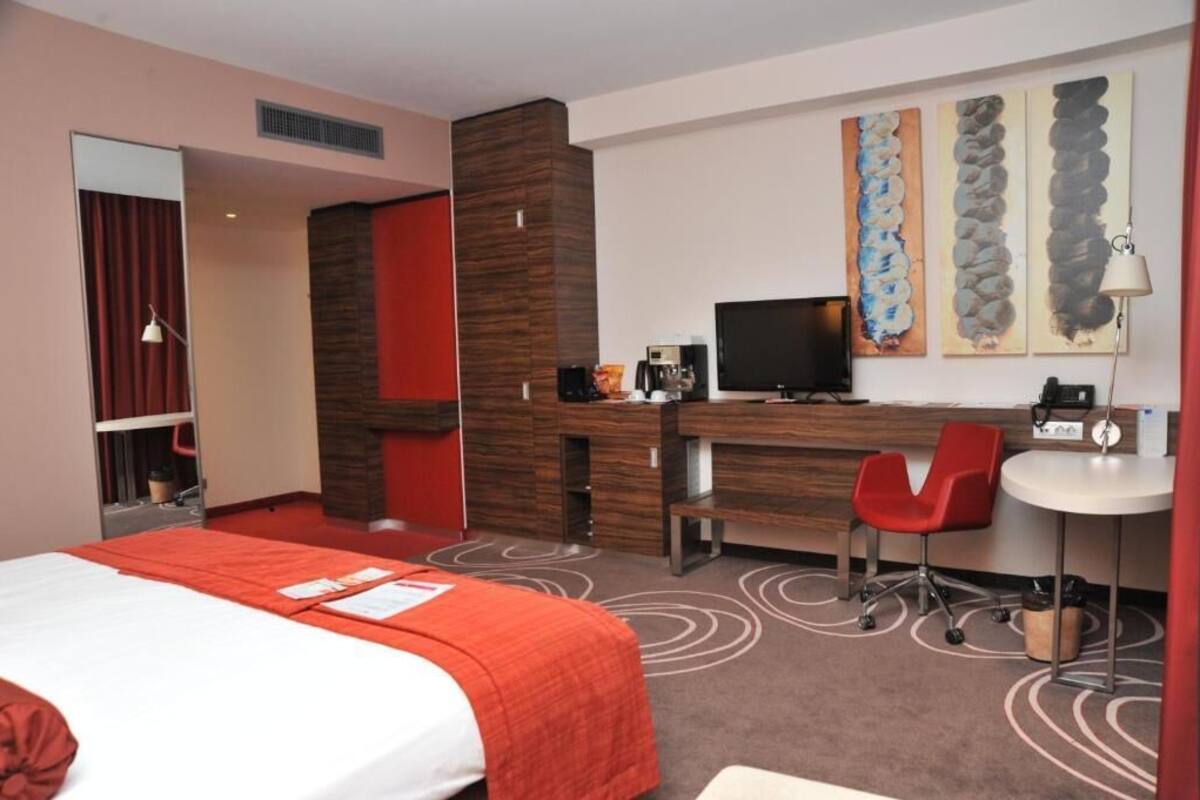 A hotel room with a bed, desk, chair, television, and artwork on the wall.
