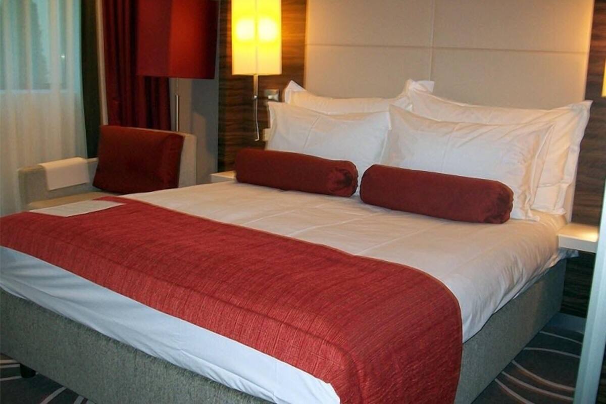 A hotel room with a large bed, red and white bedding, a red lamp, and a headboard with built-in lighting.