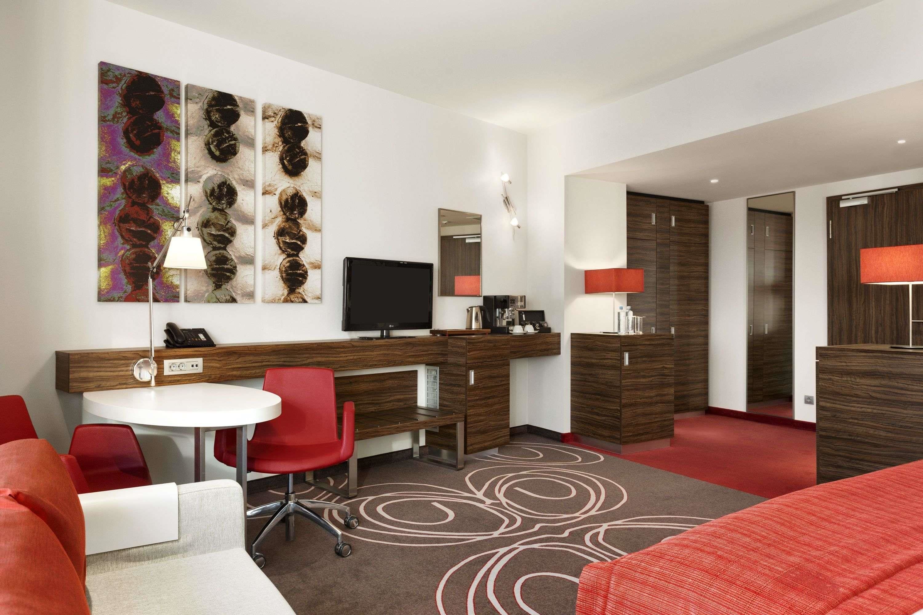 A modern hotel room with a wooden desk, red chairs, a flat-screen TV, a large mirror, and a red sofa.