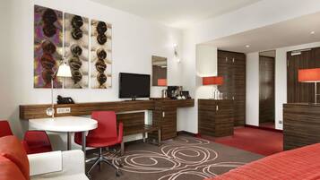 A modern hotel room with a wooden desk, red chairs, a flat-screen TV, a large mirror, and a red sofa.