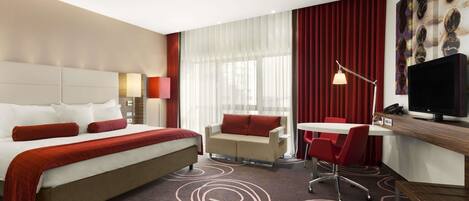 A hotel room with a large bed, a desk with a chair, a small table, and a television.