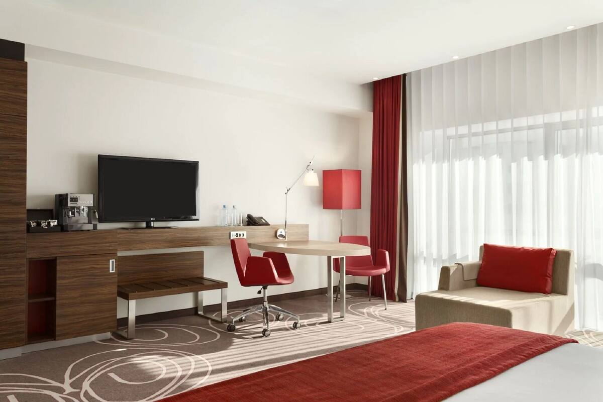 A modern hotel room with a flat-screen TV, a desk with a lamp, red chairs, a sofa with a red pillow, and a large window with sheer curtains.