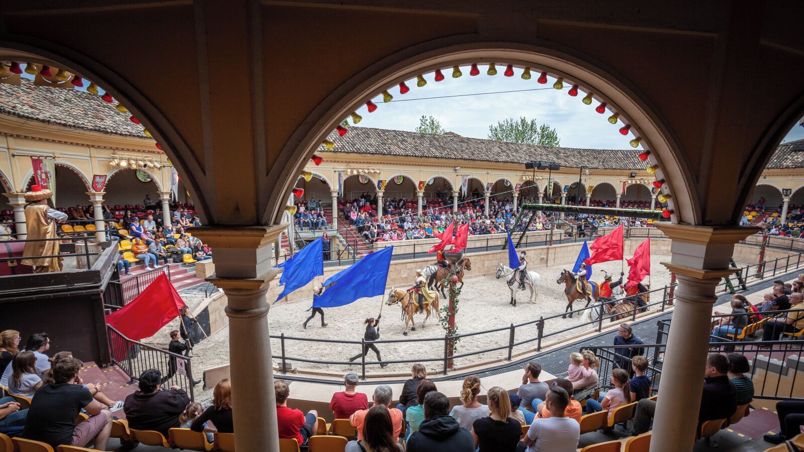 An outdoor arena with spectators, featuring a performance with horses and riders, surrounded by arches and a tiled roof.