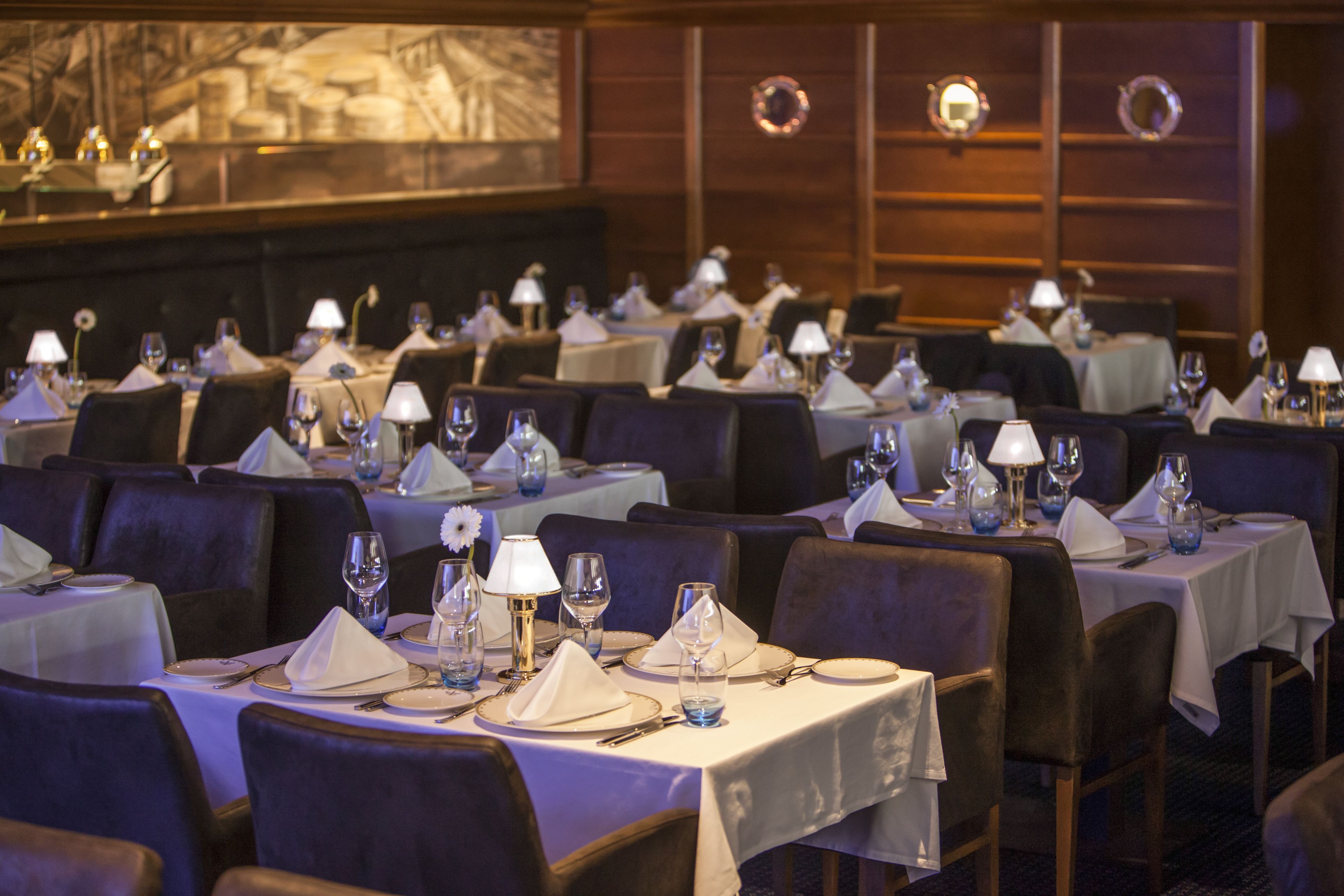 Un restaurant où de nombreuses tables sont dressées pour le service, chacune étant recouverte d’une nappe blanche et agrémentée de verres à vin et de couverts.