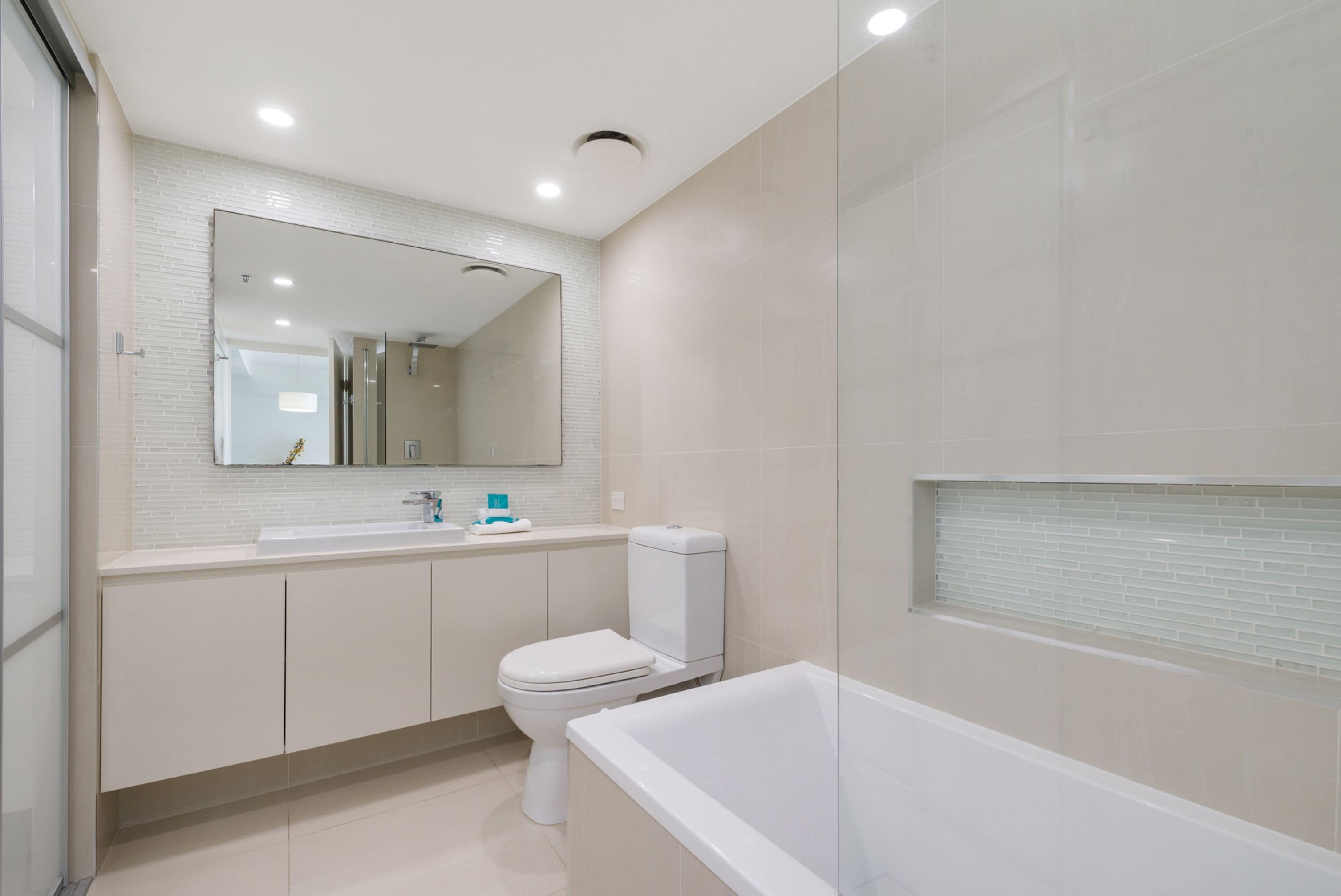 A modern bathroom with a white sink, toilet, and bathtub, a large mirror, and recessed ceiling lights.