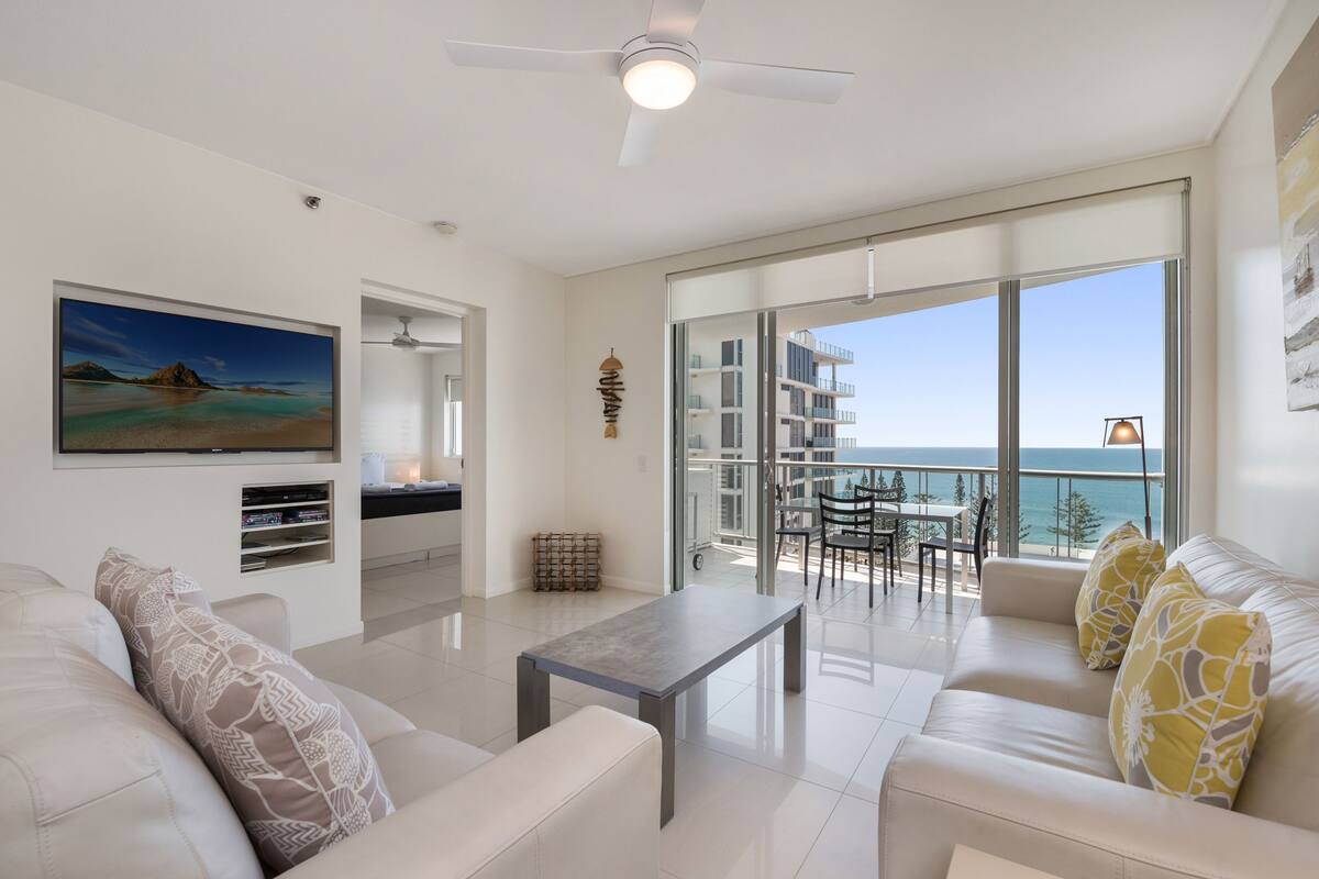 Un salon moderne avec un canapé, une table basse et un téléviseur fixé au mur. Il y a un balcon avec vue sur la mer et les bâtiments.