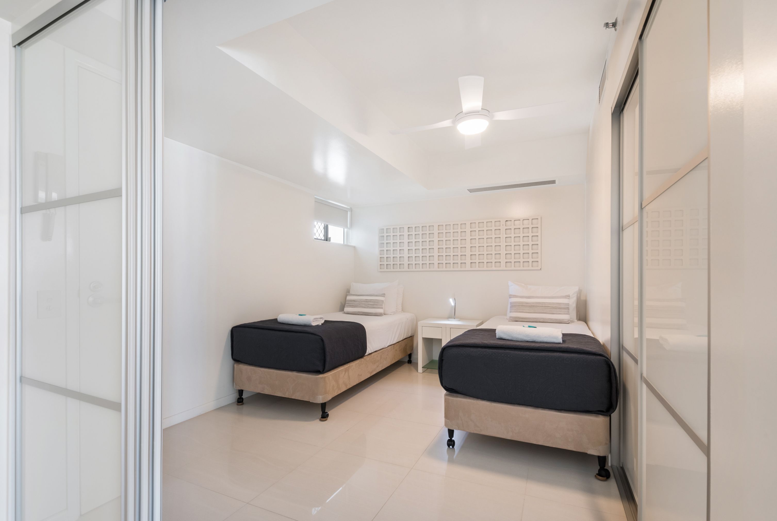 A modern hotel room with two beds, a small table, a ceiling fan, and a large window.