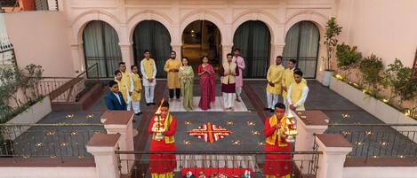 A group of people in traditional attire gathered in front of a building with a balcony, a decorative floor pattern, and a railing.