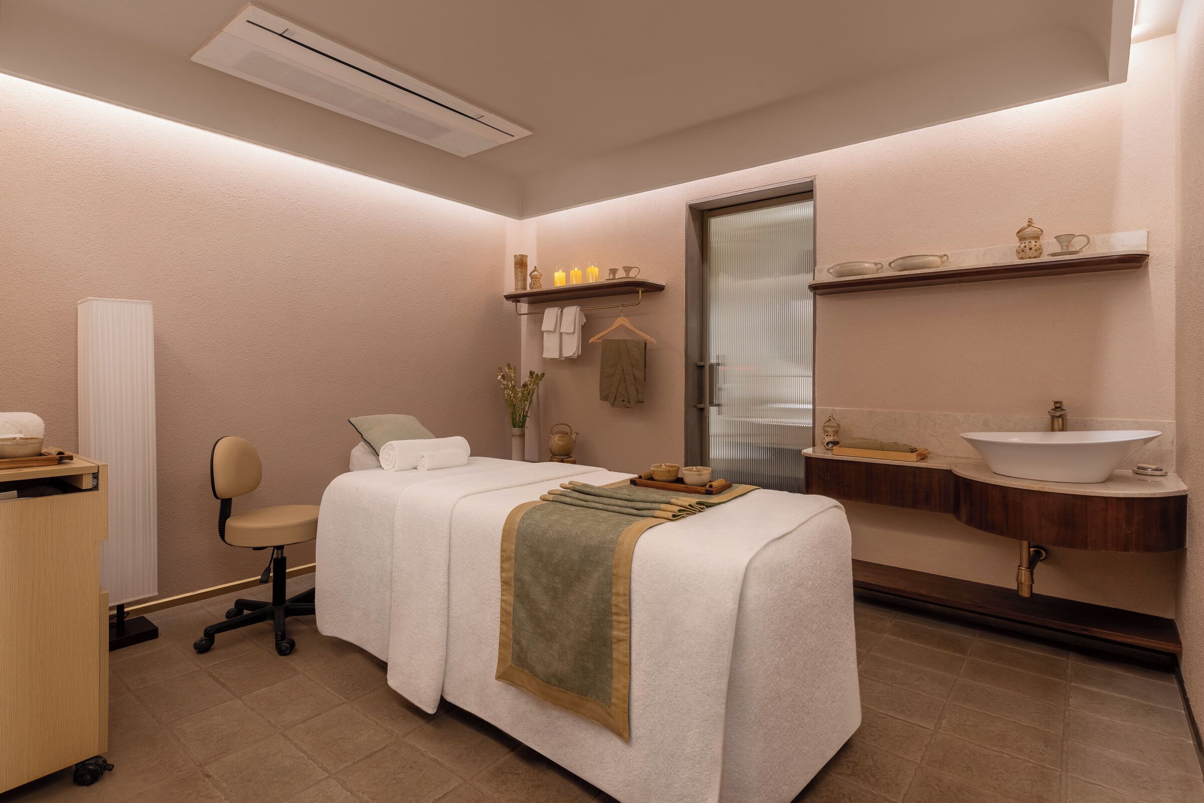 A spa room with a massage table, a small desk, a chair, a shelf with candles, and a mirror with a sink.
