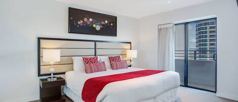 A modern hotel room with a large bed, a nightstand with a lamp, a red throw blanket, a city view through a sliding door, and a framed artwork on the wall.