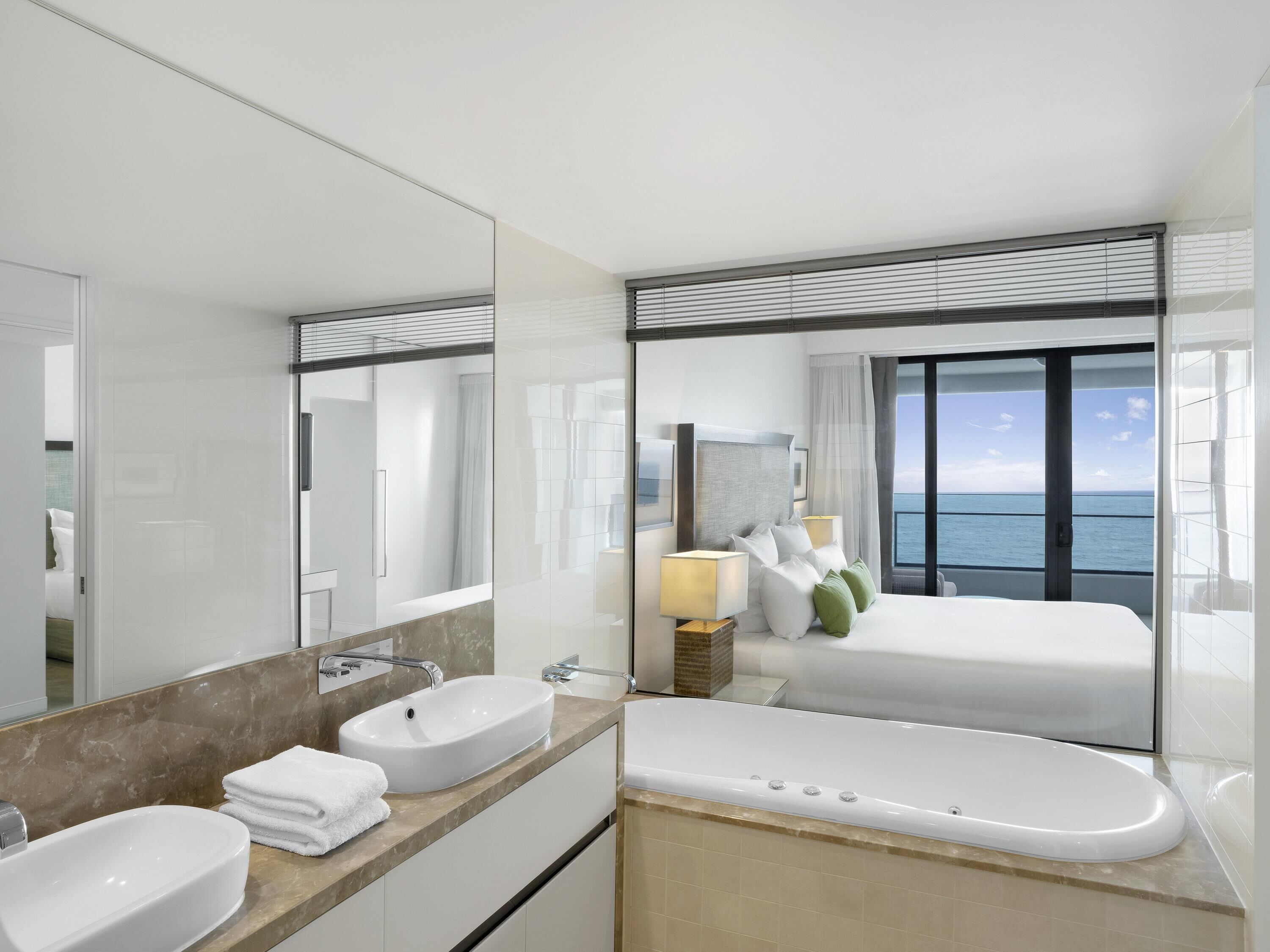 Une salle de bain d’hôtel moderne, dotée d’un grand miroir, de deux vasques, d’une baignoire et offrant une vue sur la mer.
