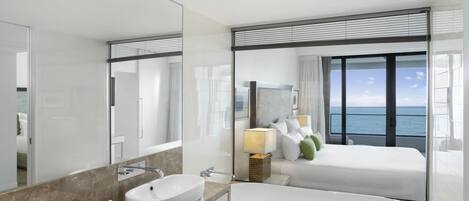 A modern hotel bathroom with a large mirror, two sinks, a bathtub, and a view of the sea.