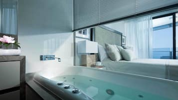 Habitación de hotel moderna con una bañera grande, una cama con almohadas, una mesita de noche con lámpara y vistas a la ciudad.