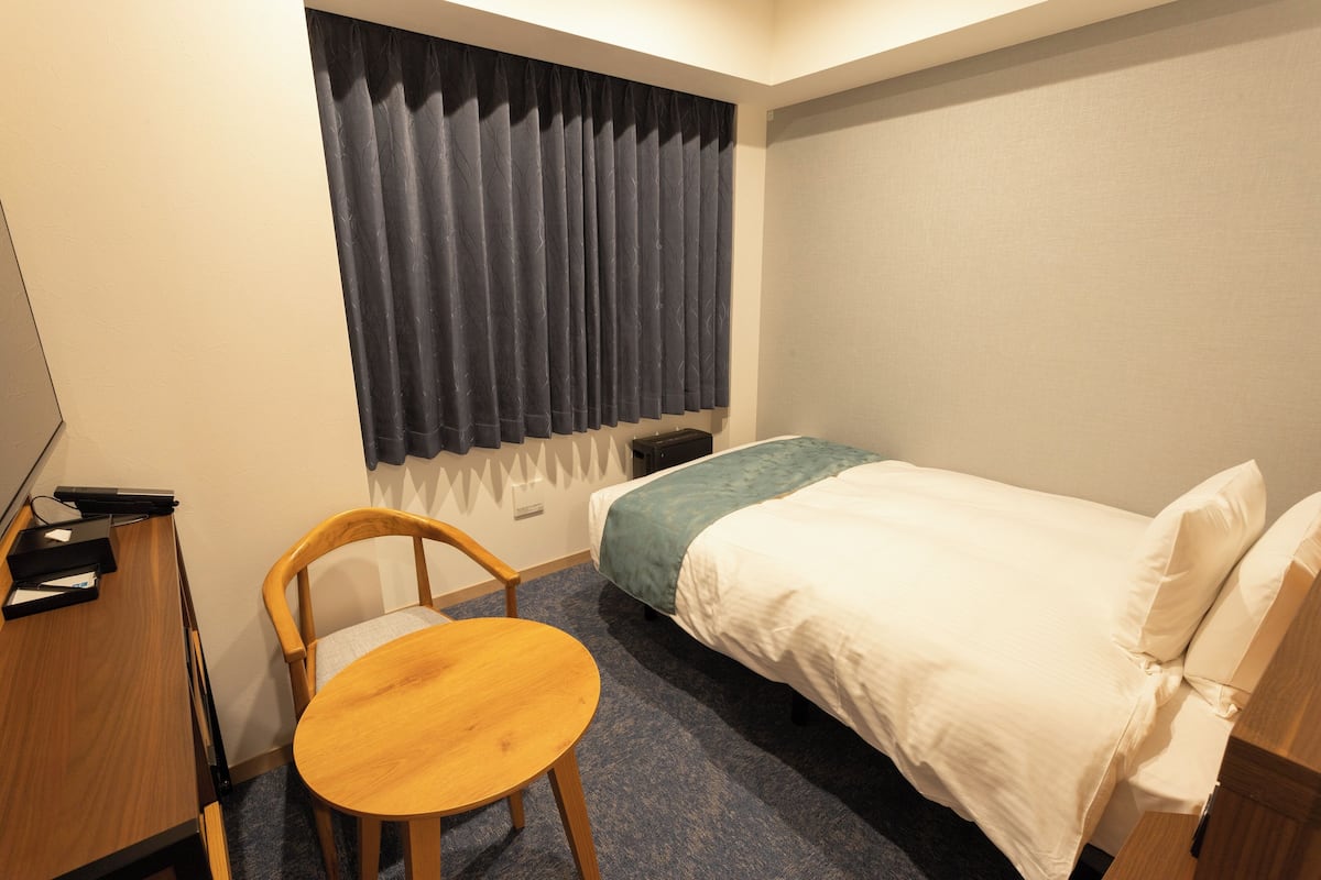 Habitación de hotel pequeña con una cama individual, una silla de madera, una mesita y un escritorio con una computadora.