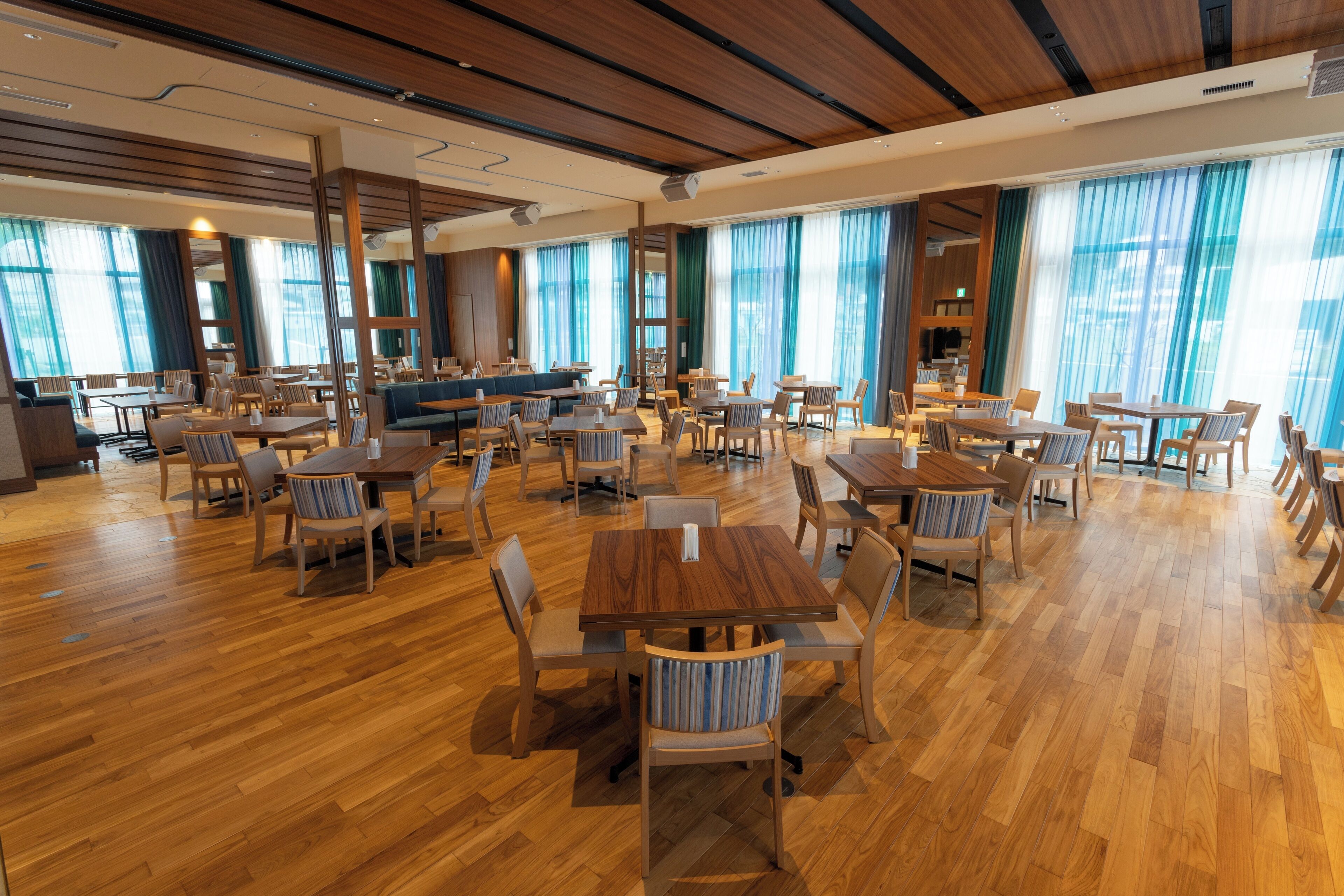 Un comedor amplio con mesas y sillas de madera, ventanas grandes con cortinas azules y un diseño de techo moderno.