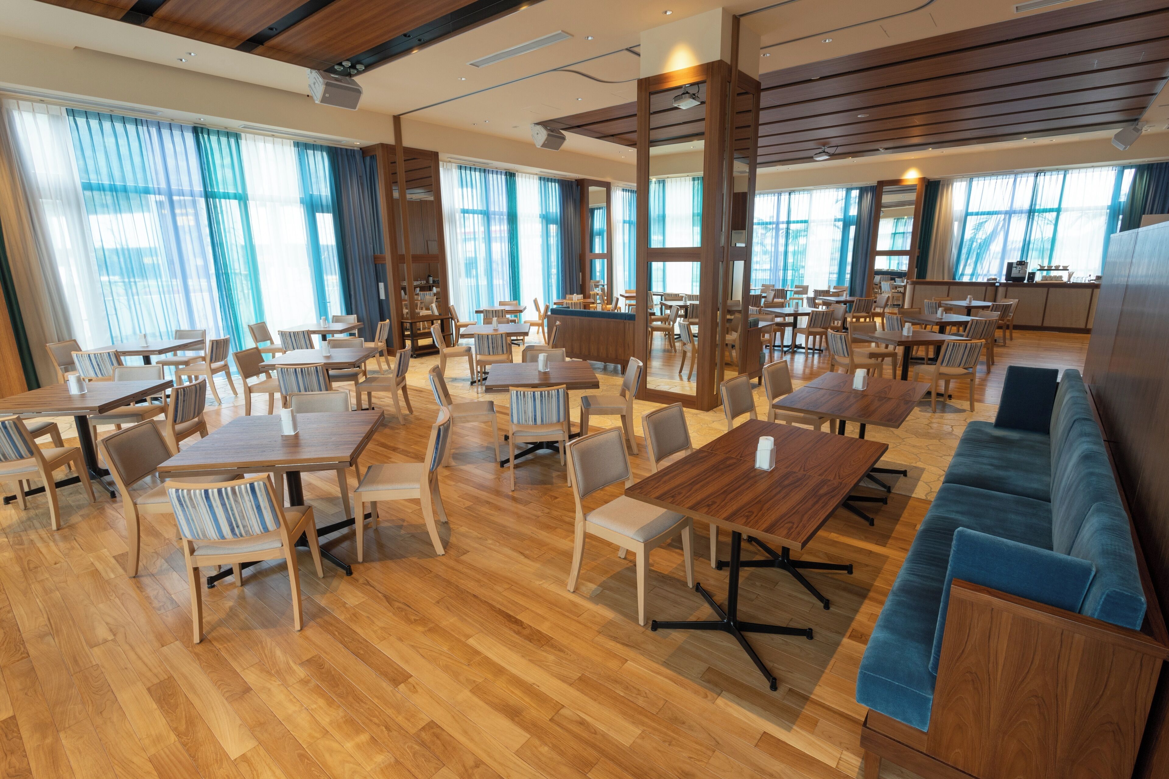 A spacious dining area with multiple wooden tables and chairs, a blue sofa, and large windows with blue curtains.