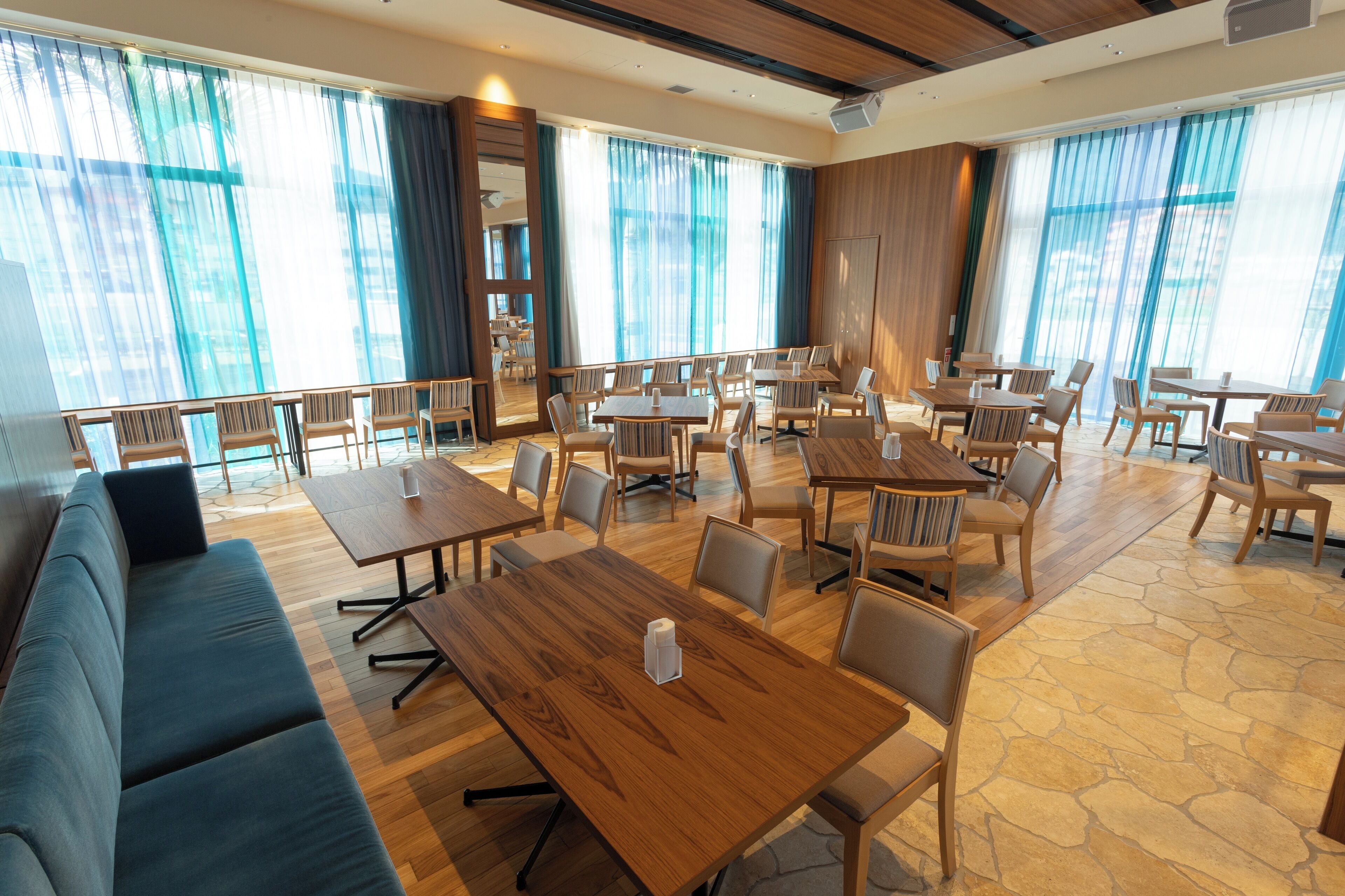 A spacious dining area with multiple wooden tables and chairs, large windows with blue curtains, and a stone-patterned floor.