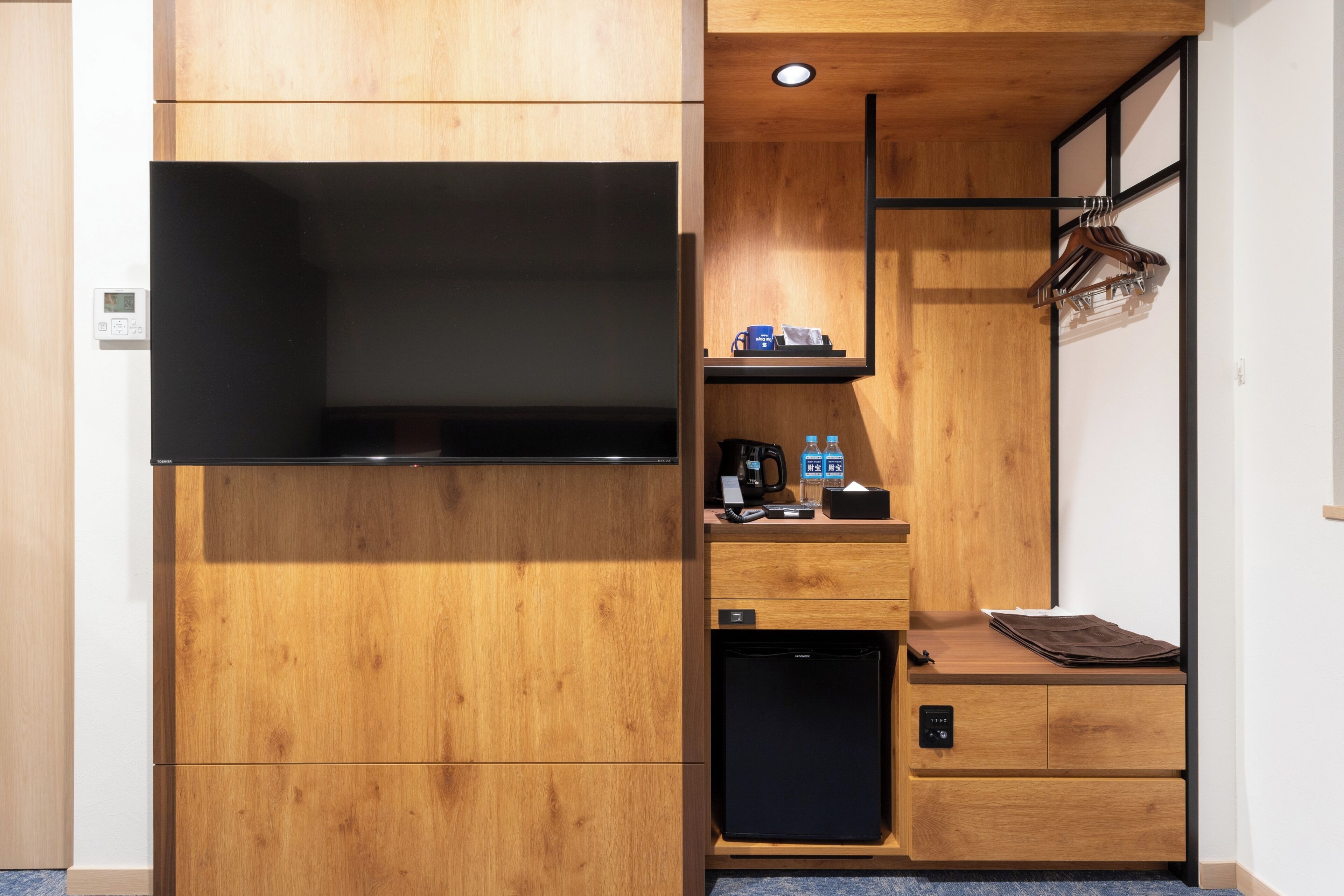 A compact room with a built-in TV, a small fridge, a coffee maker, and a hanging coat rack.