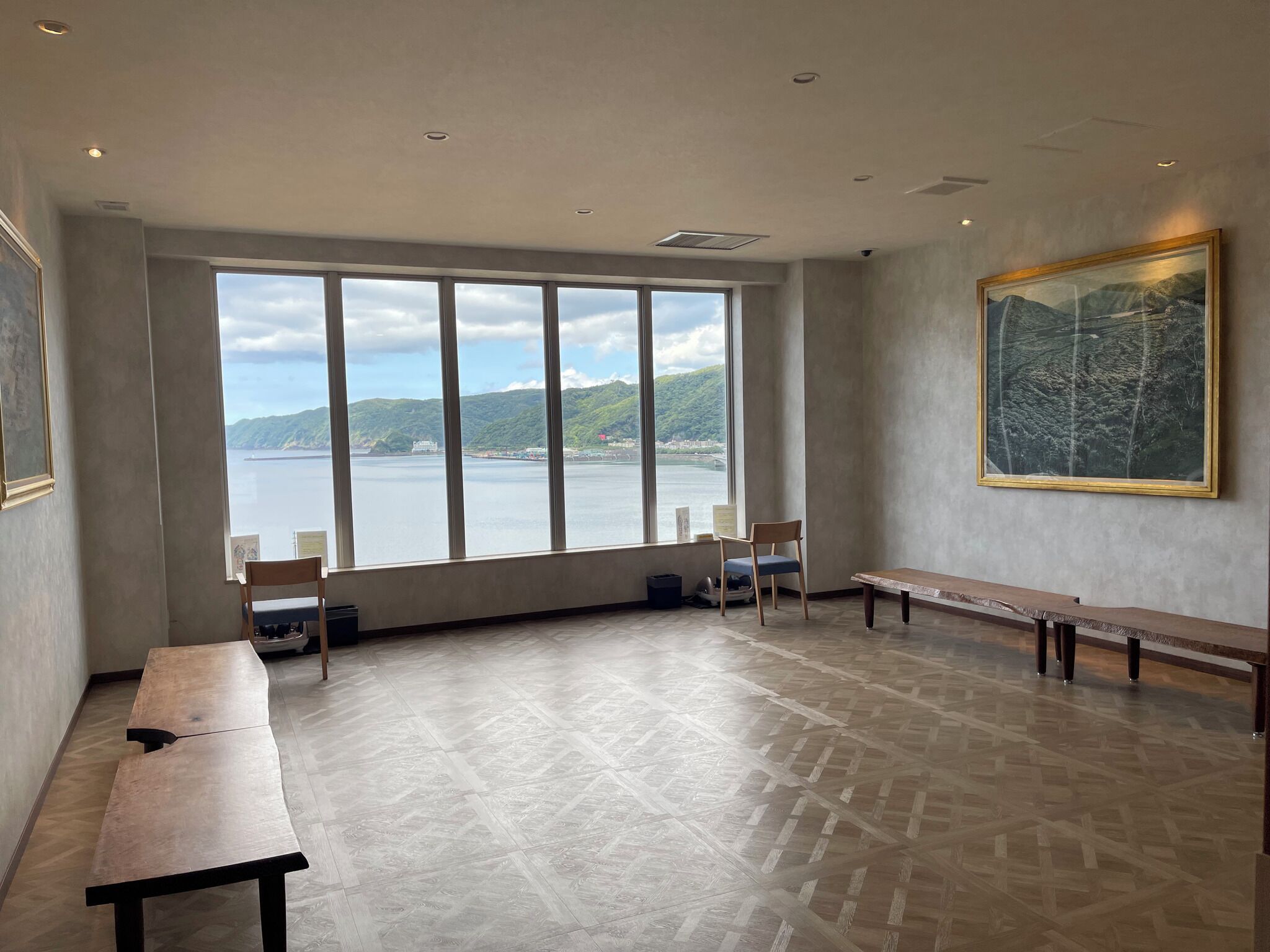Ein geräumiges Zimmer mit großen Fenstern und Blick auf ein Gewässer und Hügel, eine hölzerne Bank und ein Tisch.